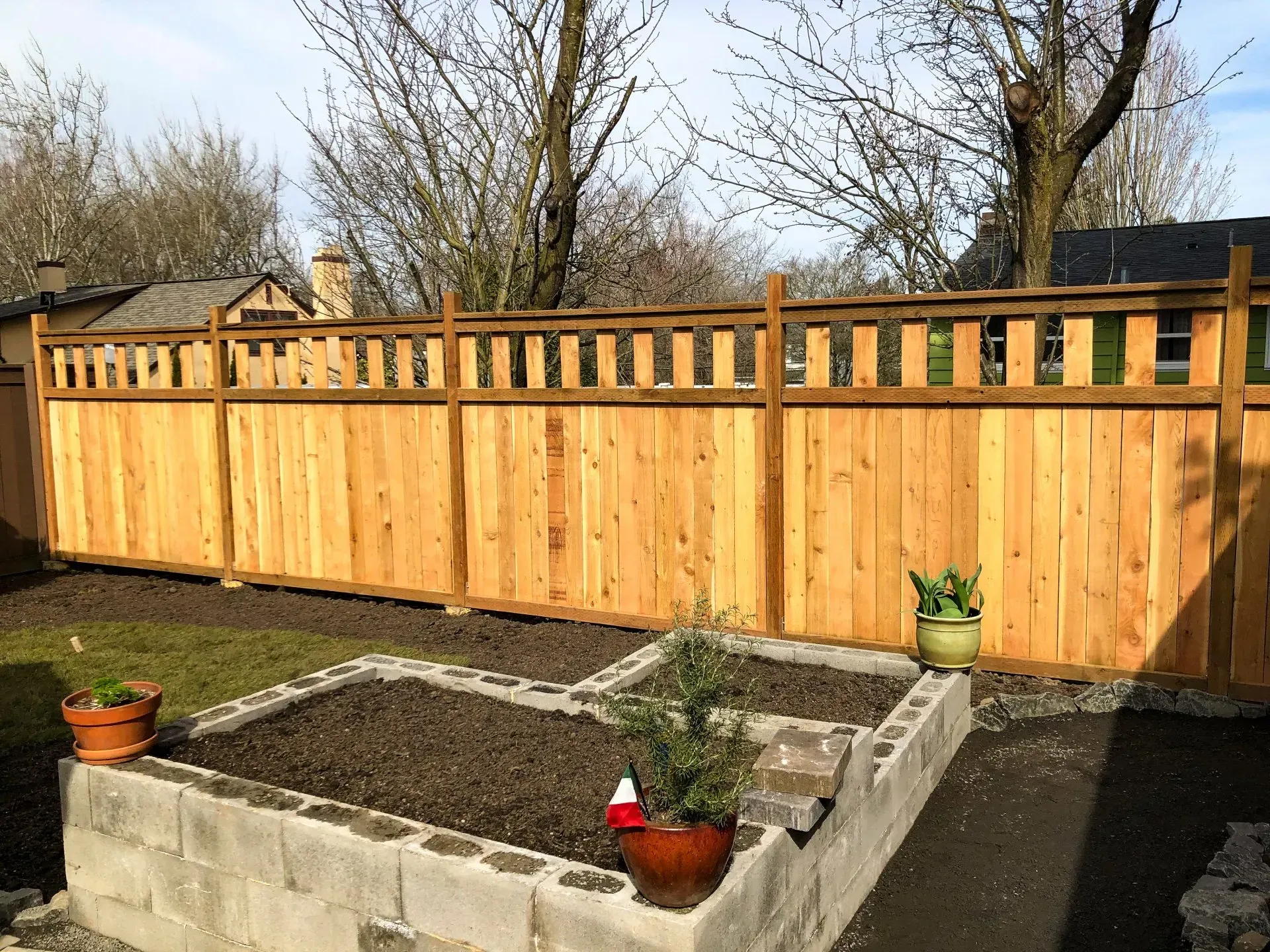 Wooden fence surrounding a backyard with a raised cinder block garden bed and potted plants.