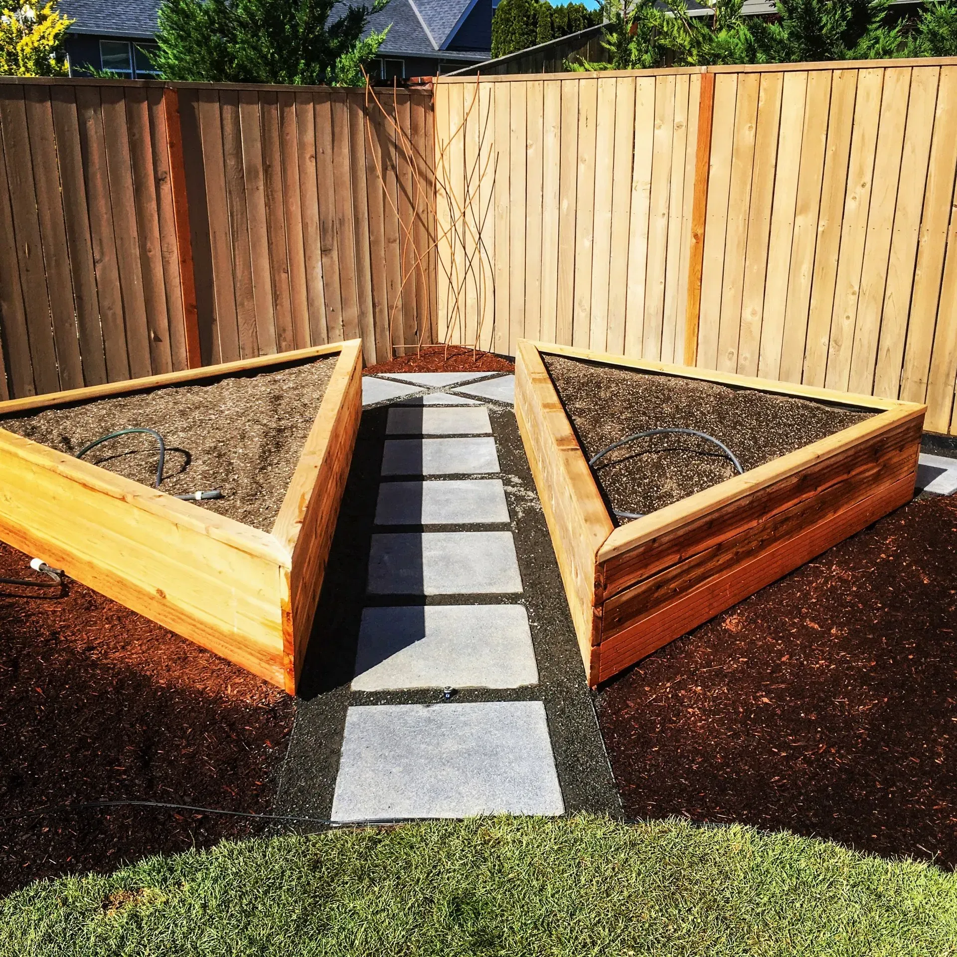 Two triangular wooden raised garden beds flank a stone walkway, set against a wooden fence in a yard.