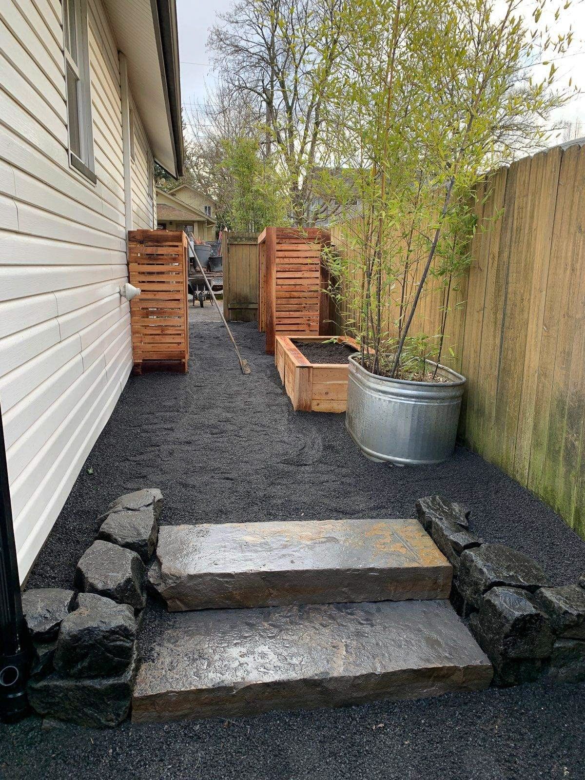 Stone steps lead to a backyard with black mulch, wooden planters, a metal container with bamboo, and a wood fence.
