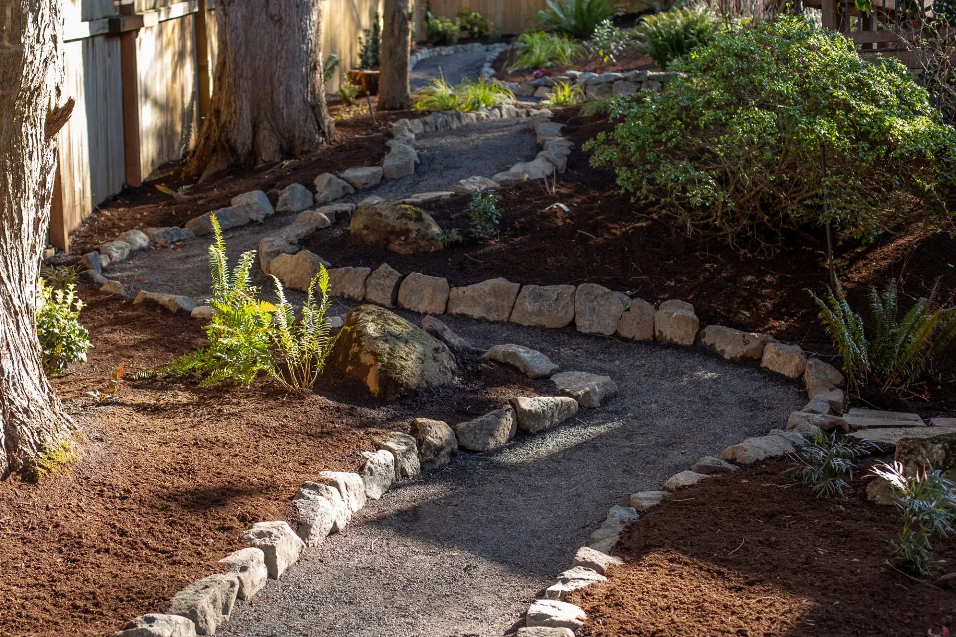 A winding gravel path edged with stones and bordered by mulch and plants in a garden. Sunlight casts shadows.