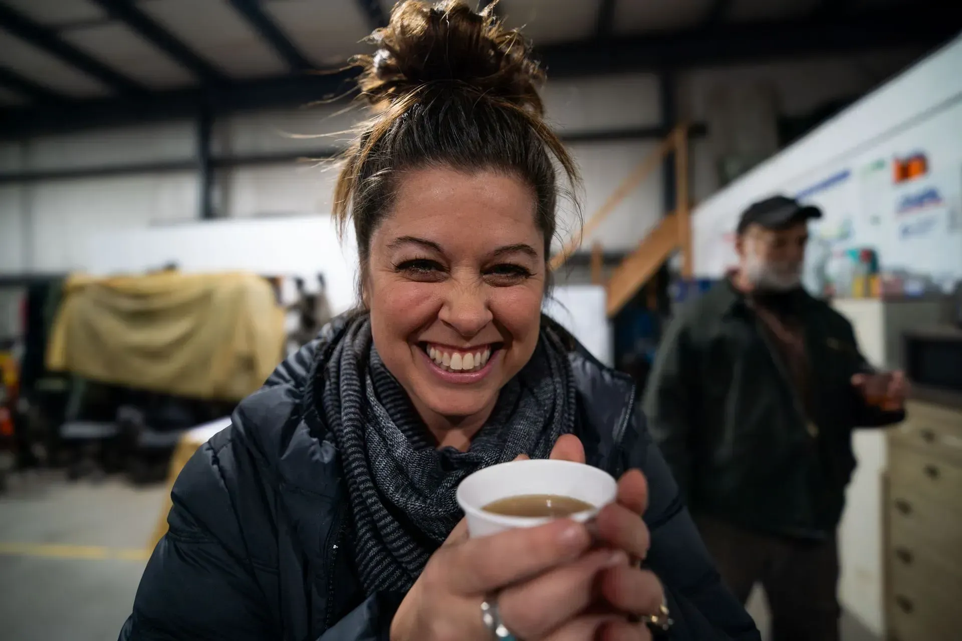 Woman with a messy bun smiles widely, holding a paper cup. 