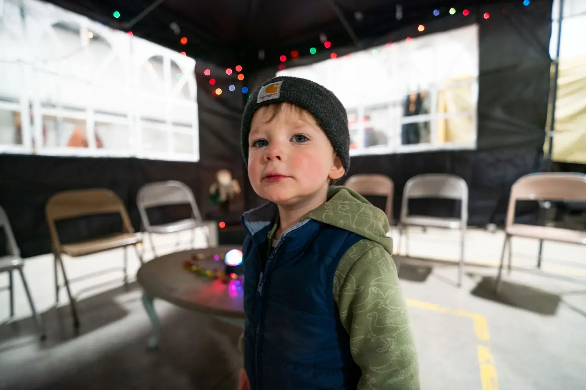 Young child in a dark room wearing a beanie and vest; chairs arranged around a small table. 
