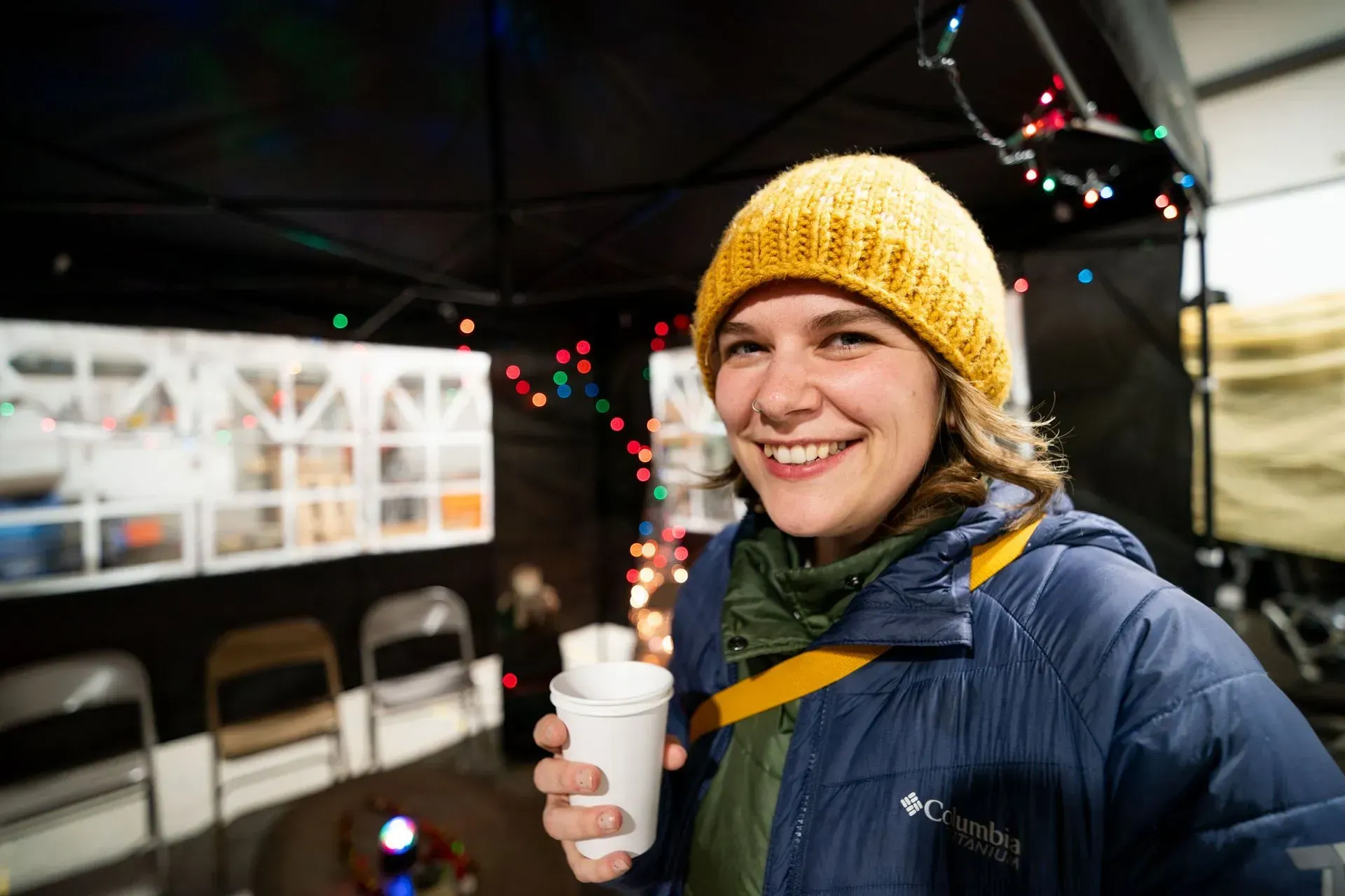 Woman wearing yellow hat and blue jacket smiles, holding a cup at an outdoor market stall with string lights.
