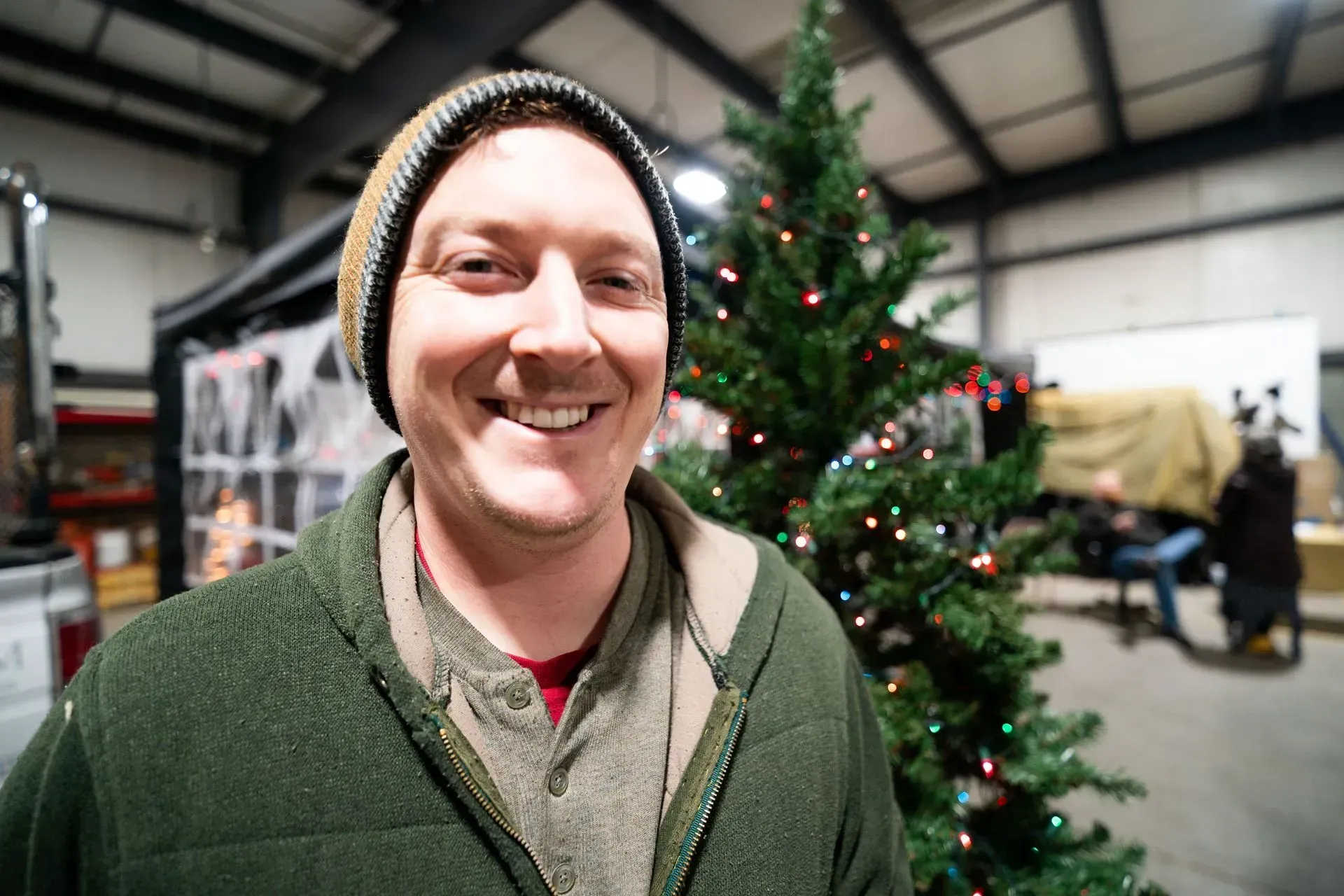 Man in beanie smiles next to a Christmas tree with lights indoors. He wears a green jacket and a red shirt.