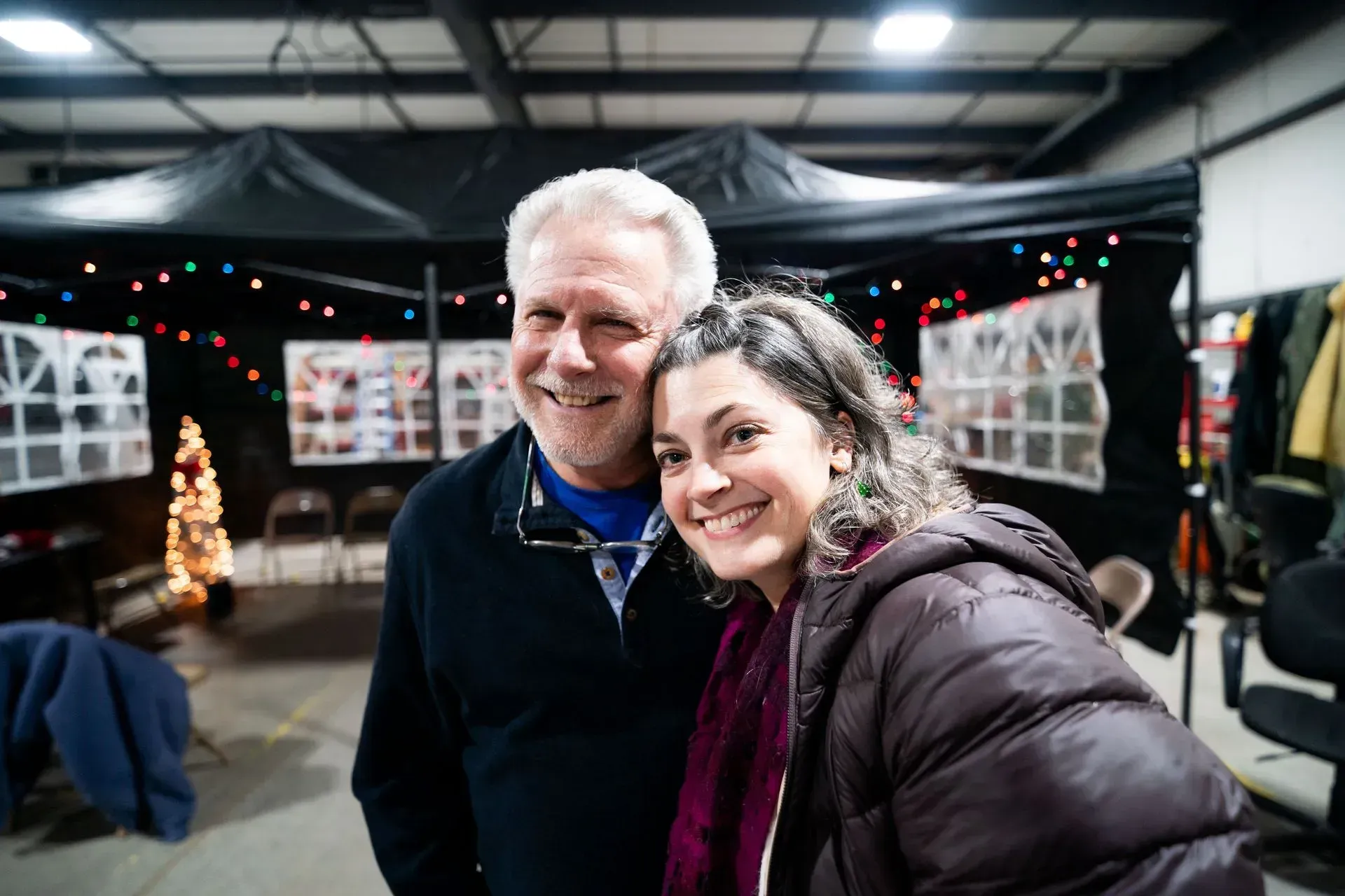 Smiling man and woman pose for a photo indoors. They are in front of a decorated space with lights and a Christmas tree.