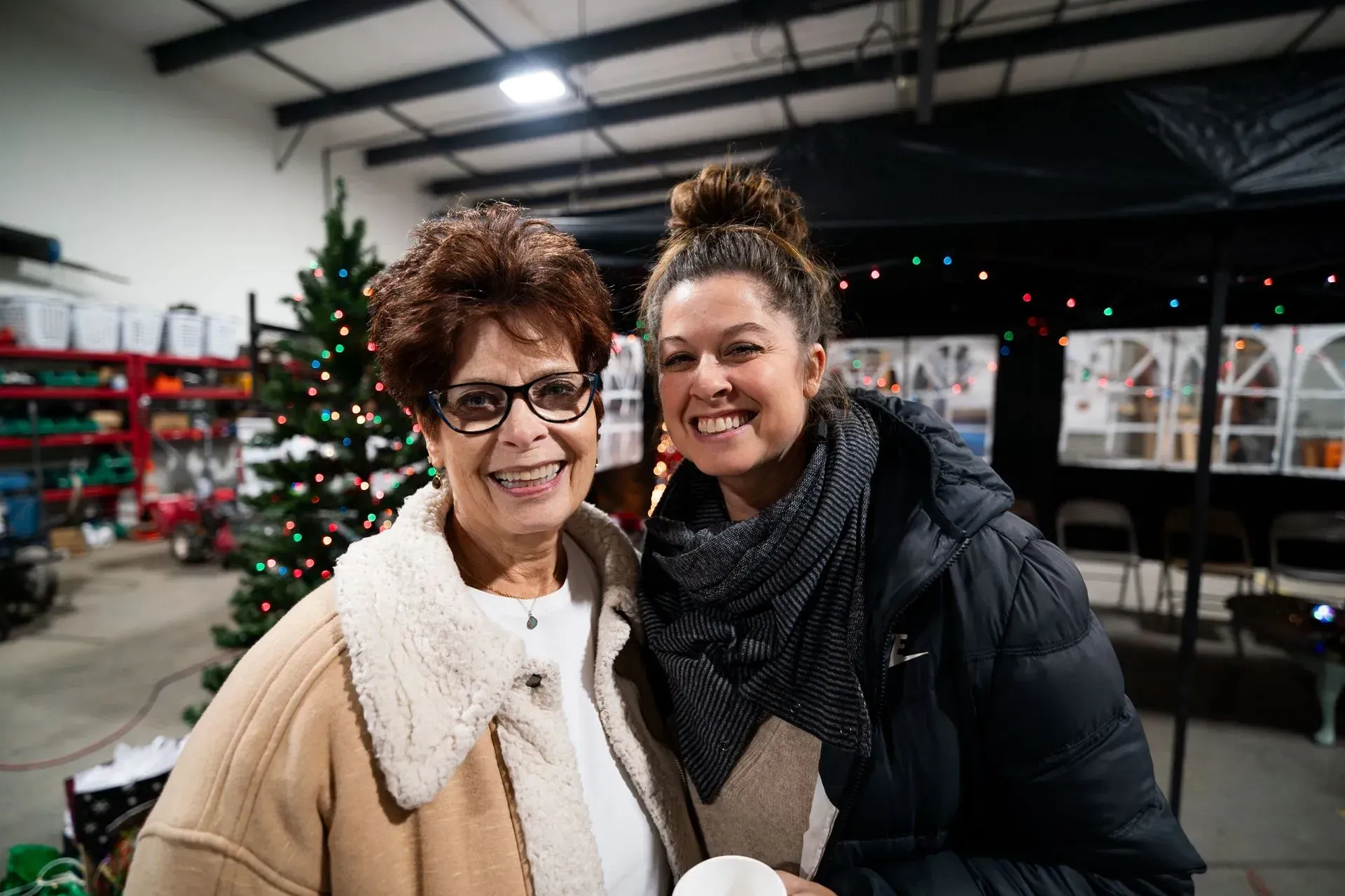Two smiling women pose together in front of a Christmas tree in a warehouse setting. 