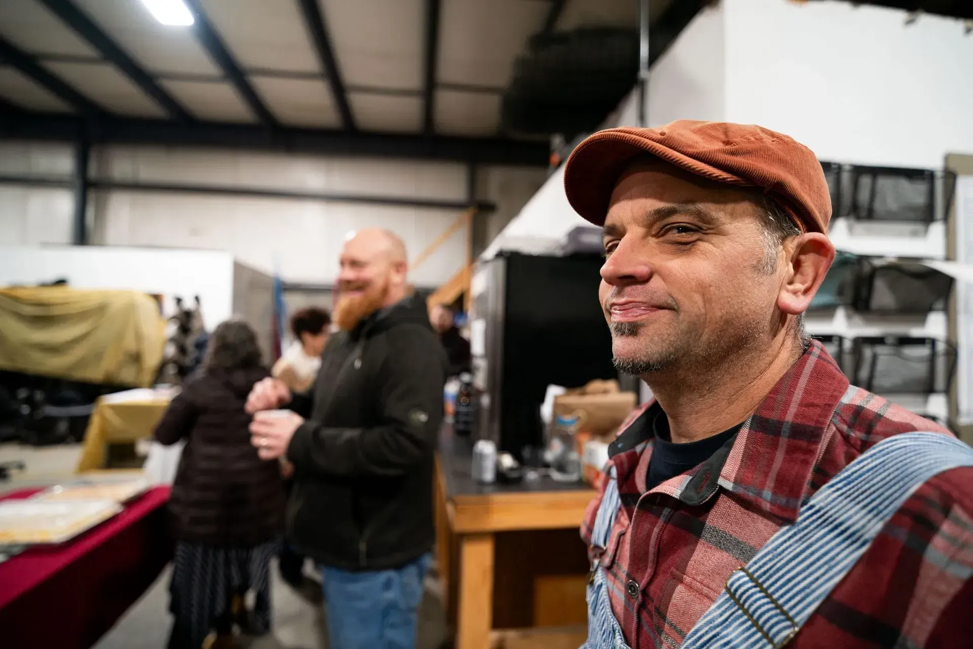 Man in a plaid shirt and cap smiles, looking right, at an event. 