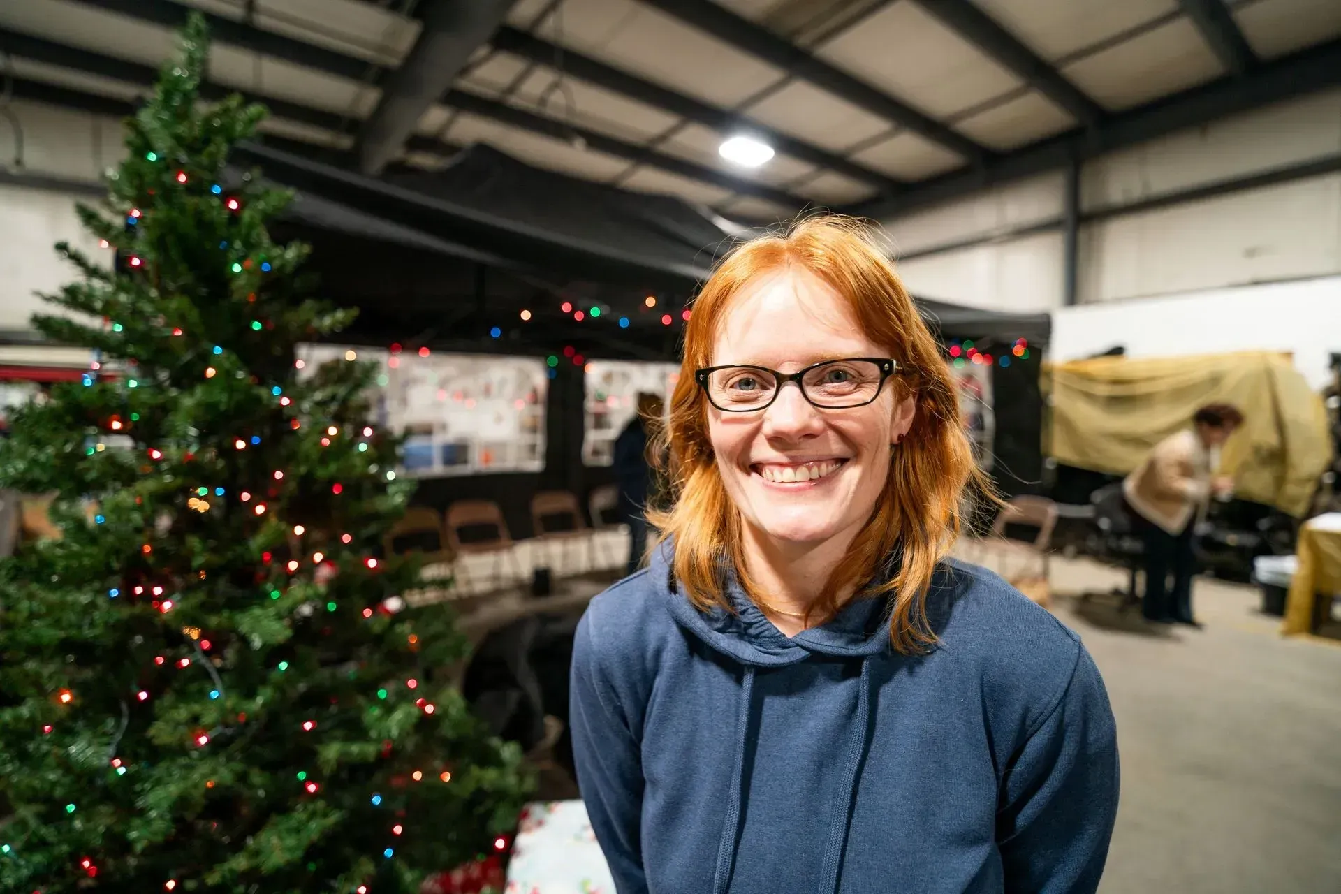 Woman with red hair and glasses smiles in front of a Christmas tree lit with colored lights. 