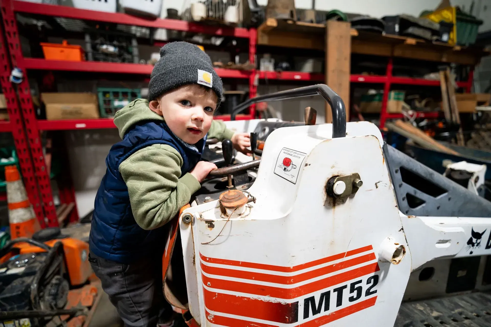 A young child wearing a beanie and vest sits on a Bobcat MT52 skid steer loader in a workshop, looking at the camera.