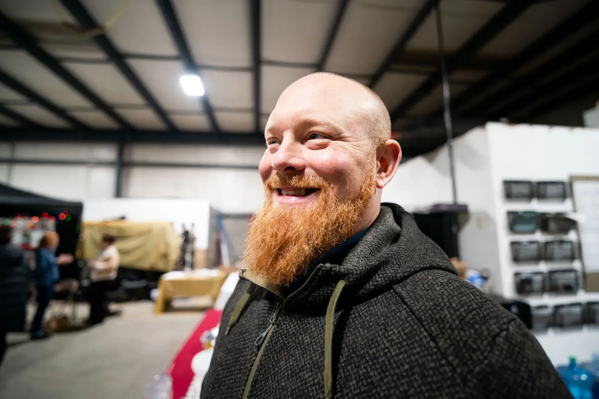 A smiling man with a red beard and shaved head in a warehouse setting; wearing a gray hooded jacket.