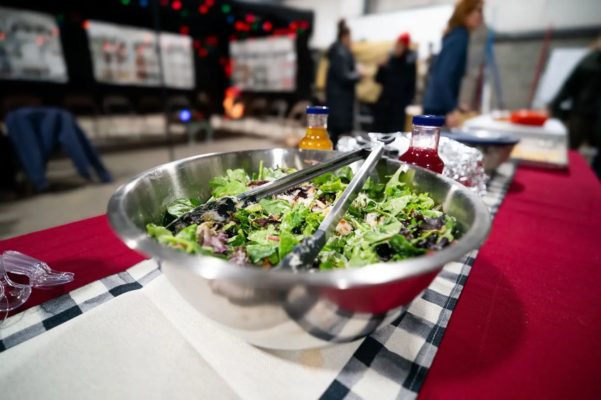 Large salad bowl on a table with two bottles of juice and people in the background.