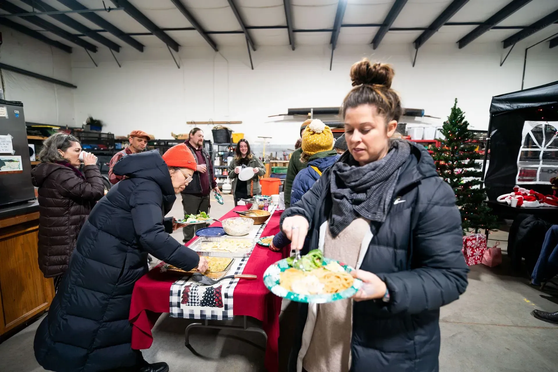 People serve themselves food from a buffet at an indoor gathering.  A woman in a dark coat holds a plate of food.