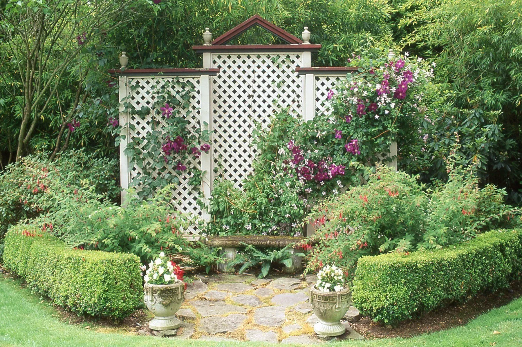 A garden with a lattice backdrop, climbing flowers, and a stone bench surrounded by hedges and potted plants.