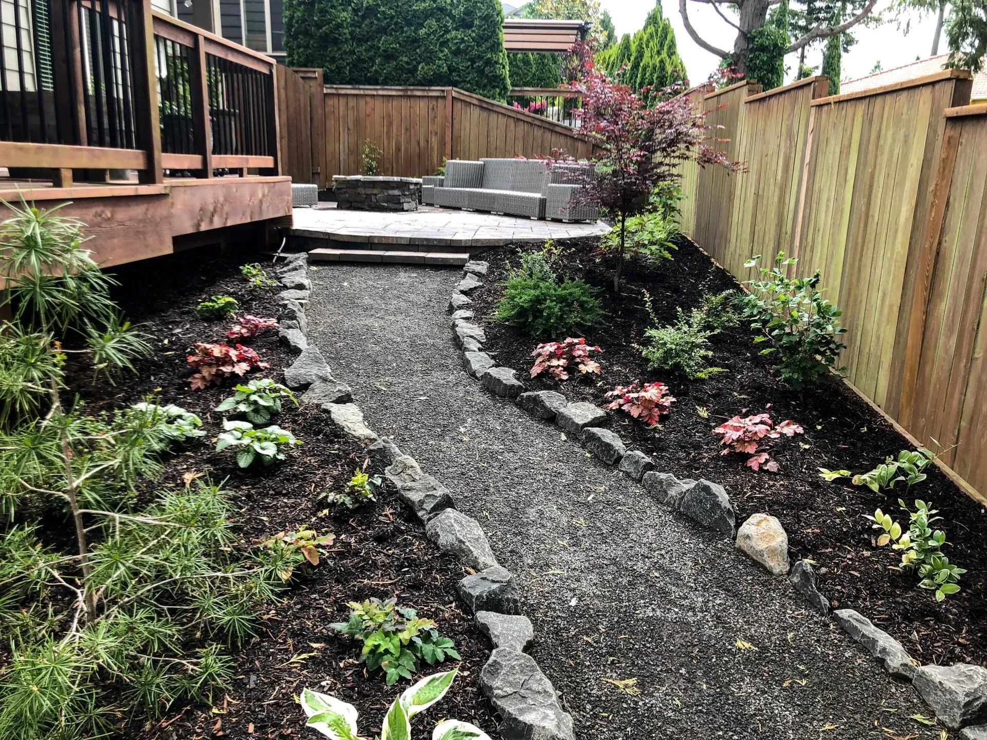 A landscaped garden with a stone path, leading to a fire pit area with a wooden deck and a wooden fence.