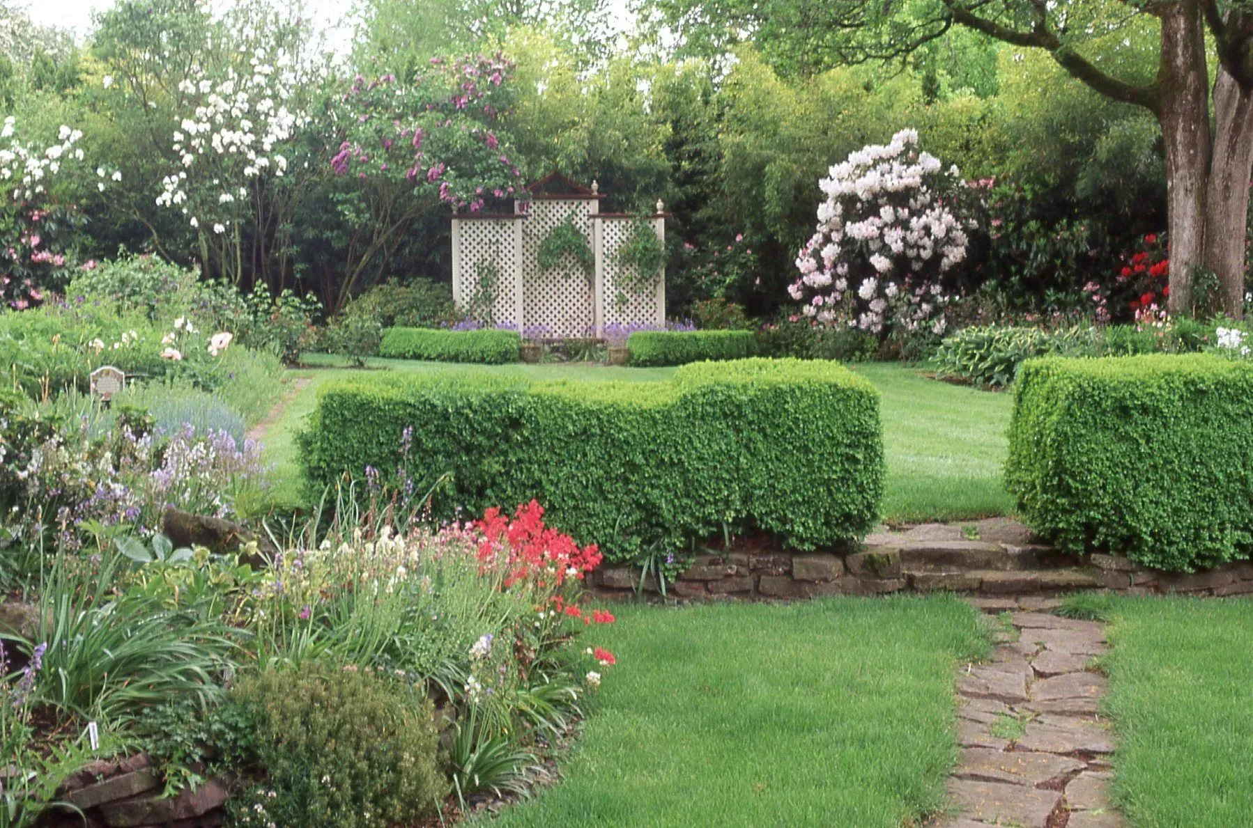 A lush garden scene featuring manicured hedges, a stone path, and flowering plants under a canopy of trees.
