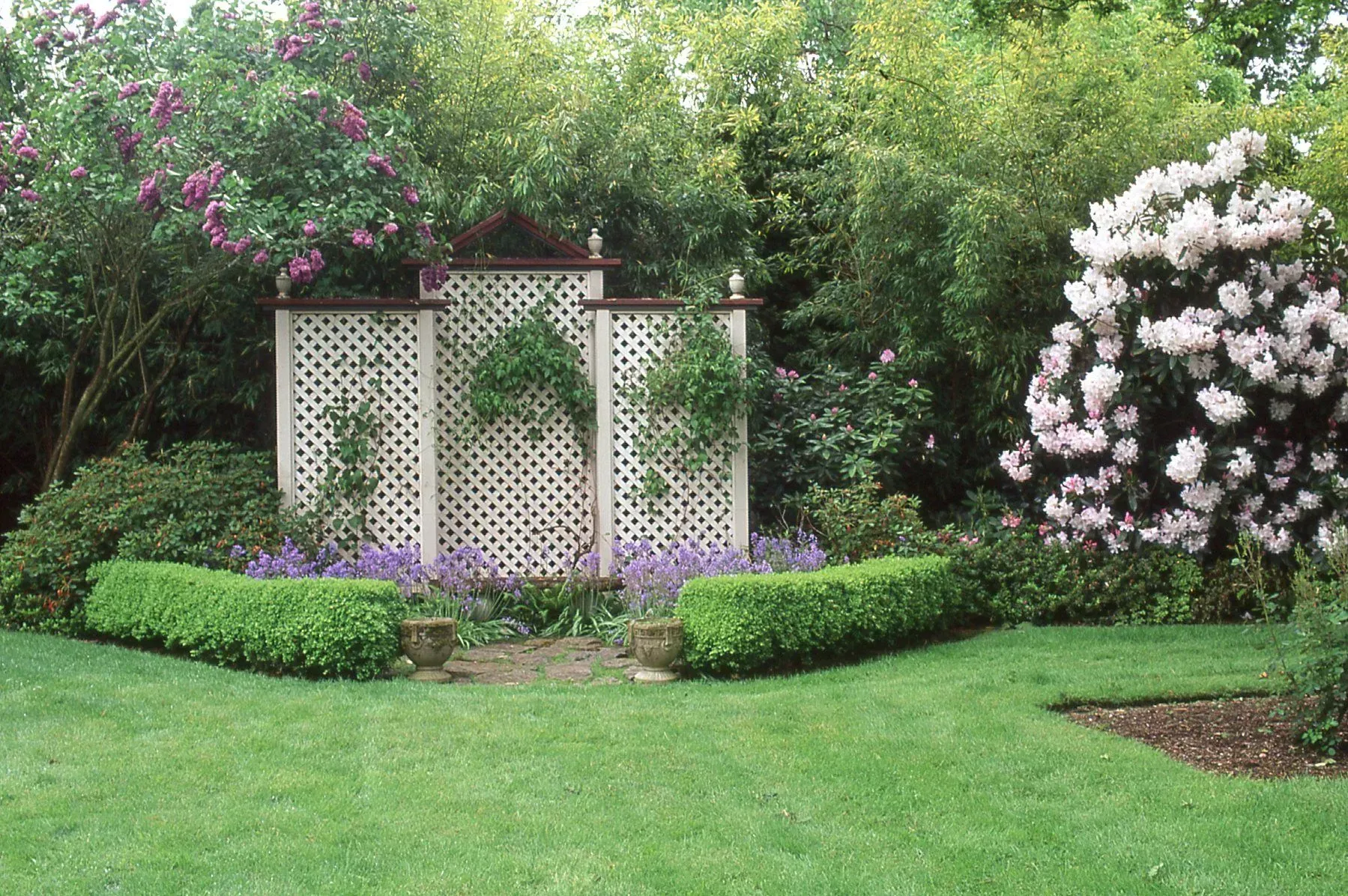 Lush garden scene with green lawn, box hedges, and a decorative trellis flanked by flowering shrubs and trees.