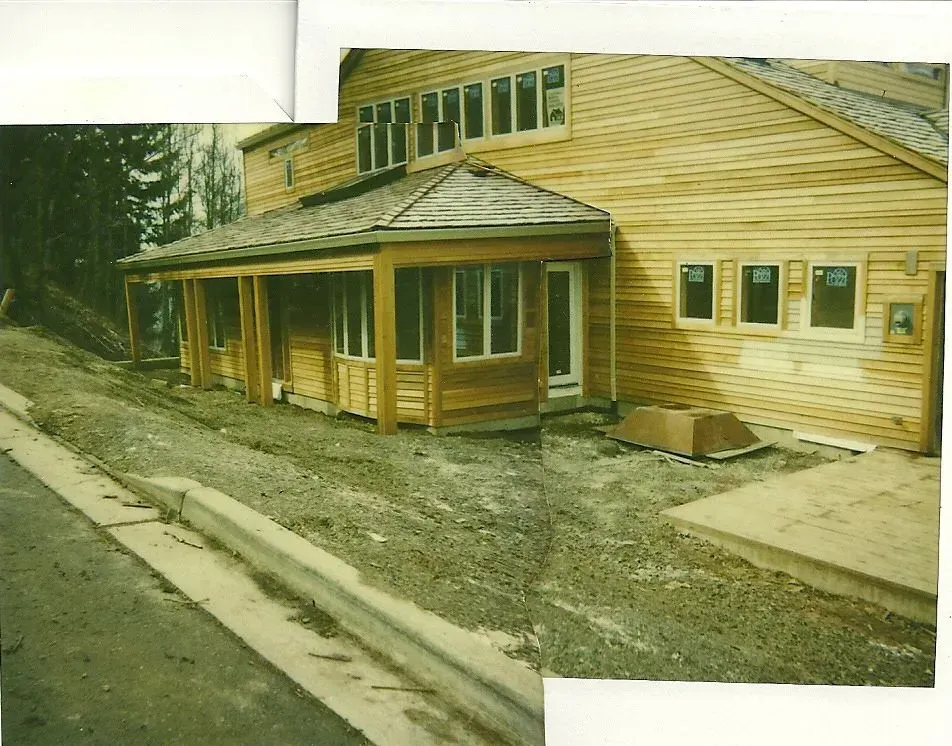 A wood-sided building under construction with a covered porch. Gravel and a concrete curb are in the foreground.