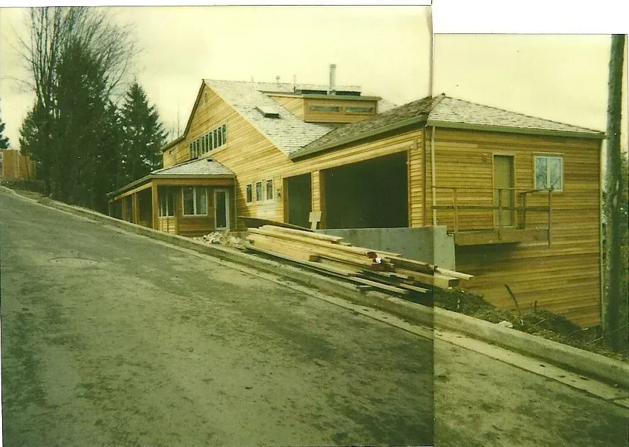 A wooden house under construction on a sloped street. Lumber and building materials are visible near the foundation.