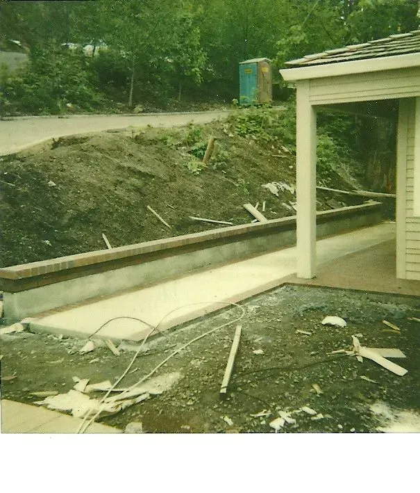 A concrete pathway under construction next to a building with a covered entrance. A portable toilet sits in the background.