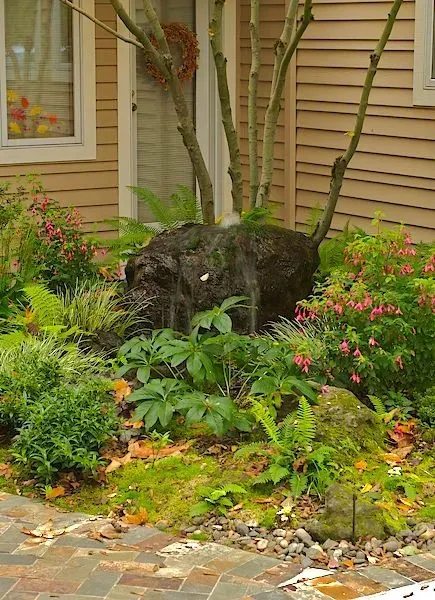 A lush garden bed with diverse greenery surrounds a tree trunk against a tan house. Pink flowers and moss add color.
