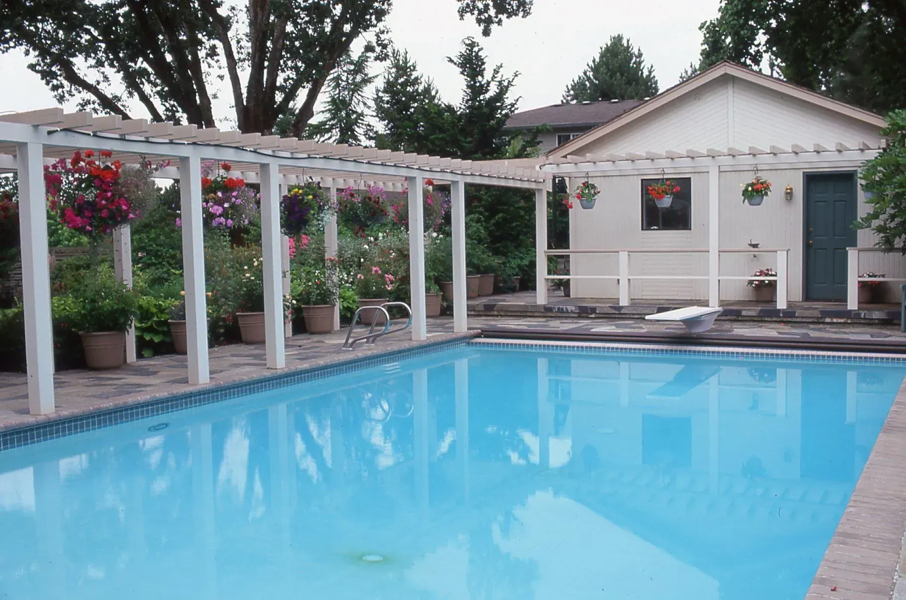 Swimming pool with a white pergola and flowers; a small building is in the background.