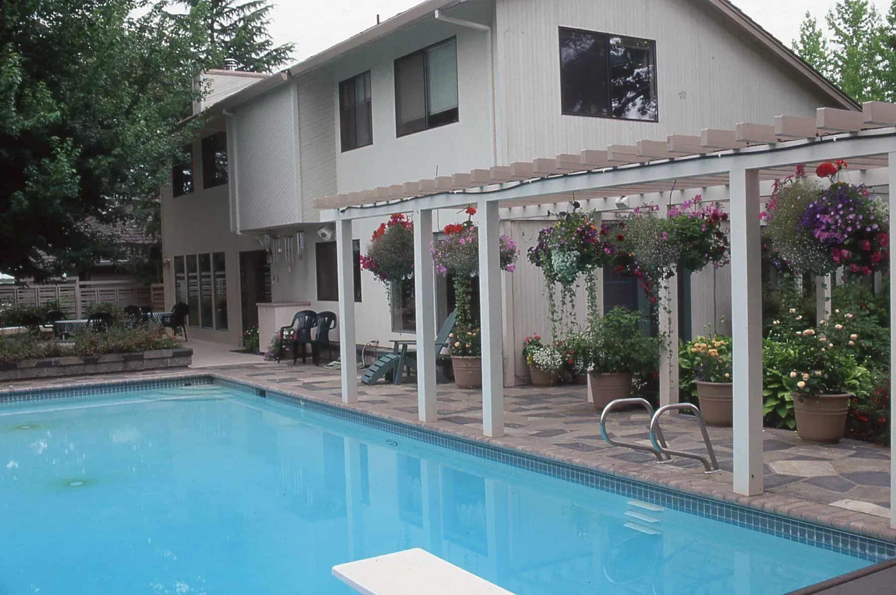 Backyard with a pool, patio, and two-story house. Hanging flower baskets adorn a pergola by the pool.