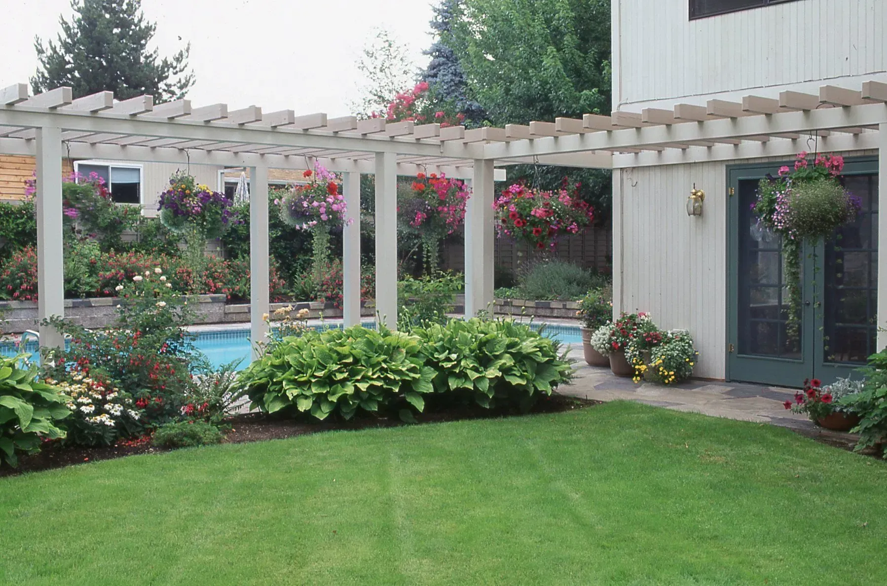 A backyard with a pool, a pergola, and lush gardens. The area is surrounded by green grass and a light-colored house.
