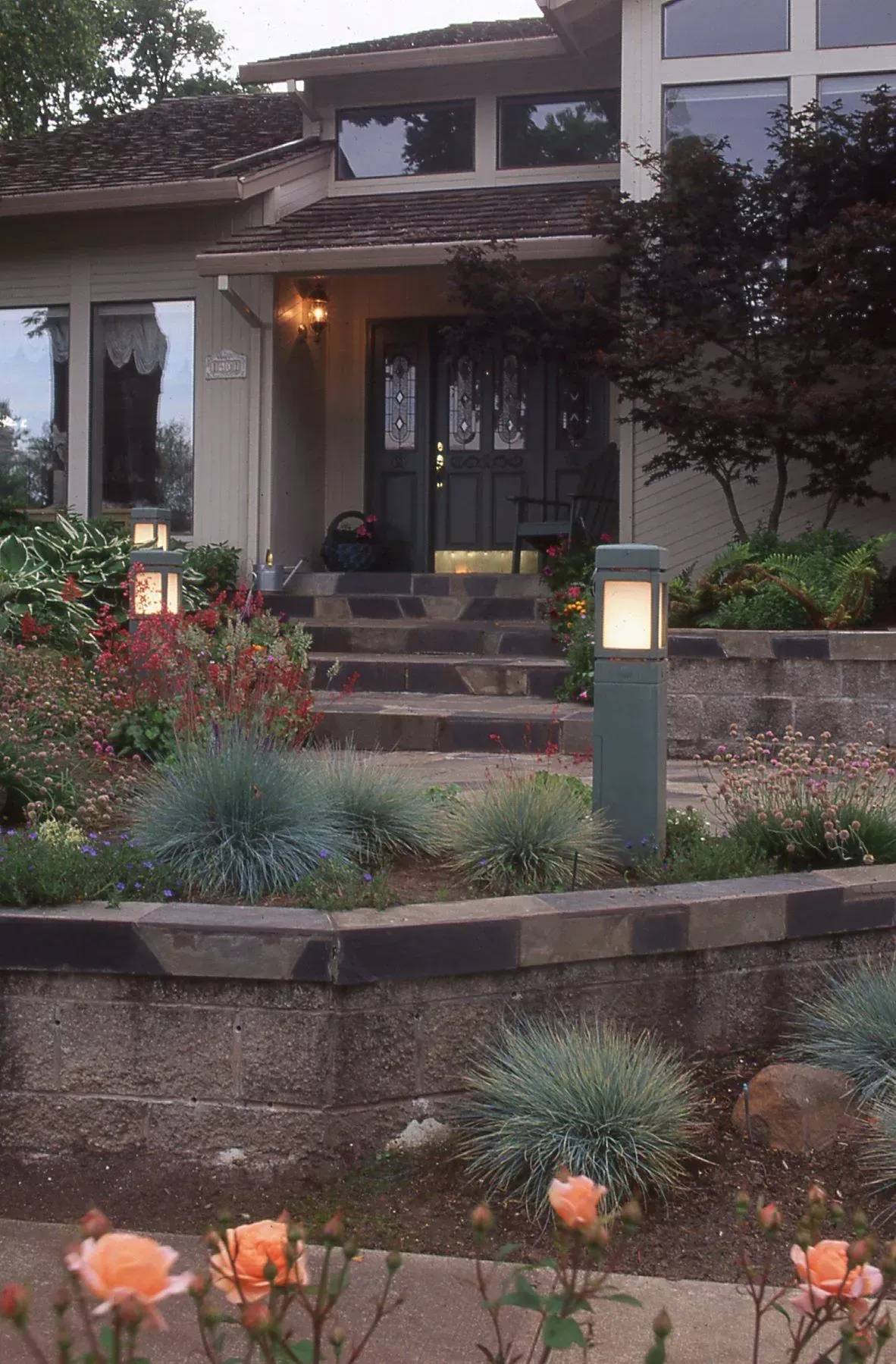 A house entrance with tiered landscaping. Gray stone wall with blue-green ornamental grasses and orange roses.