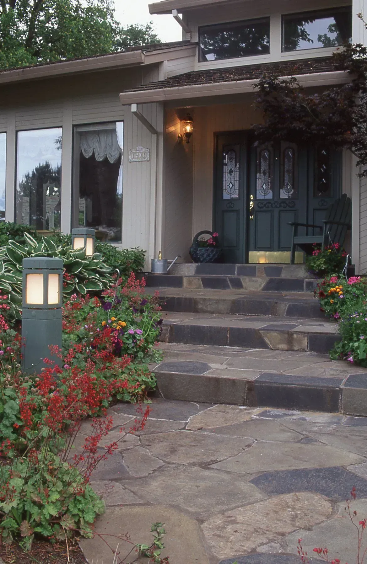 A house with a stone path leading to the front door, lined with colorful flowers and illuminated by modern outdoor lights.