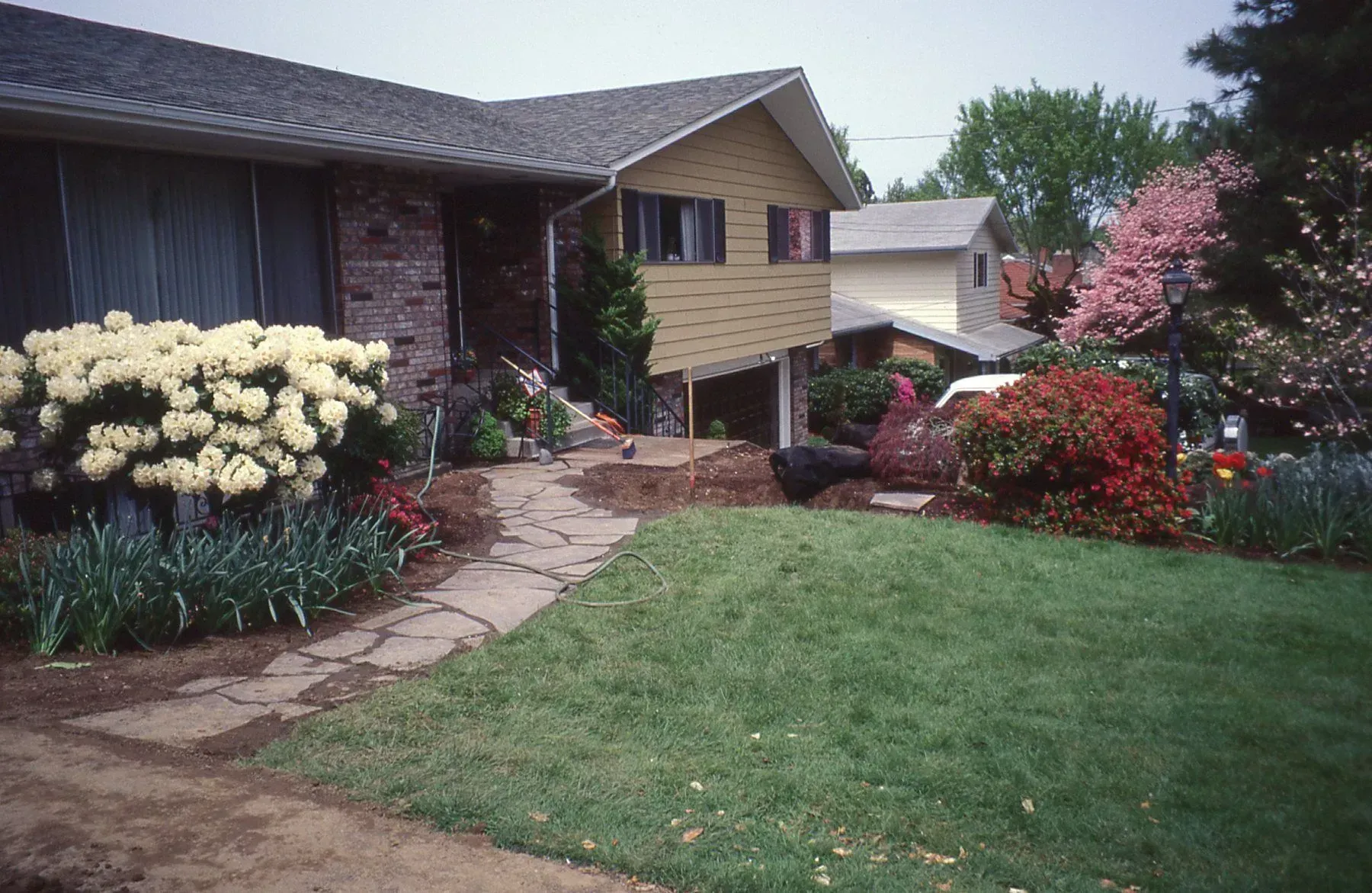 A house with a well-landscaped front yard featuring a stone walkway, various colorful flowers, and lush green grass.