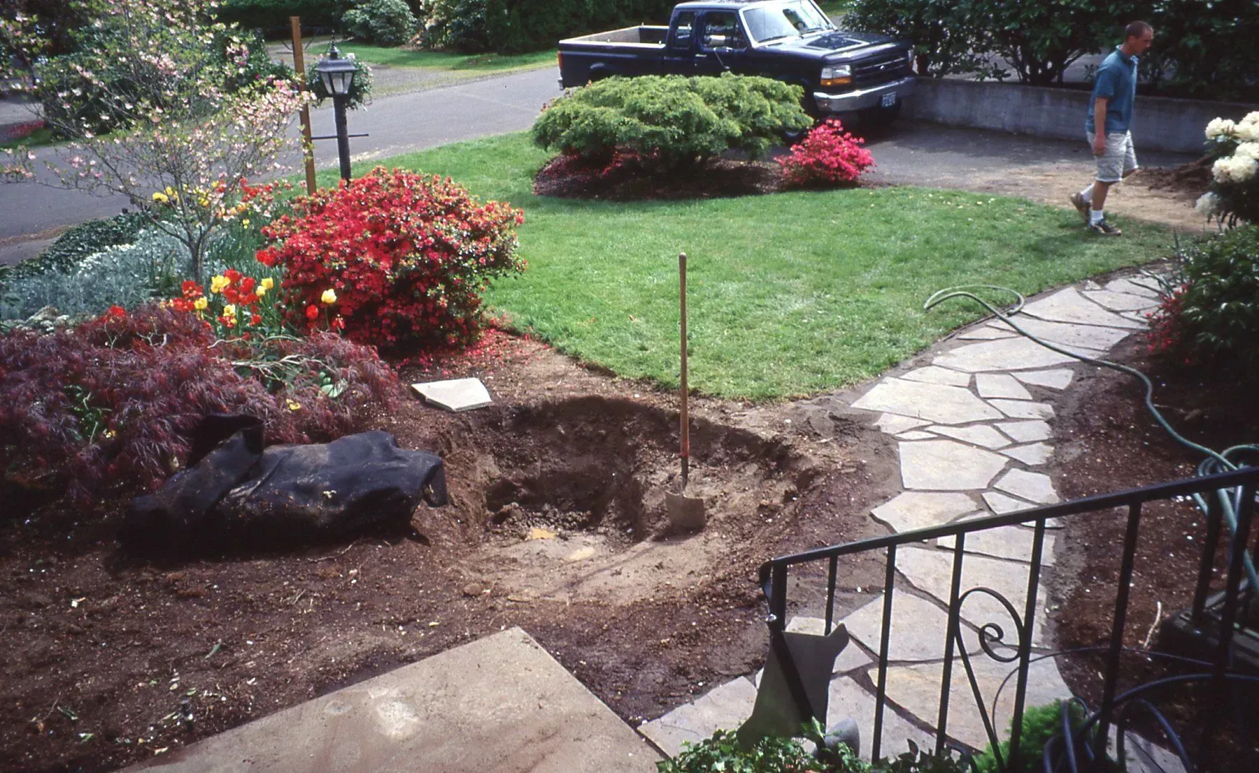 A person stands near a front yard with a freshly dug hole, surrounded by a stone path, lawn, flowers, and a truck.