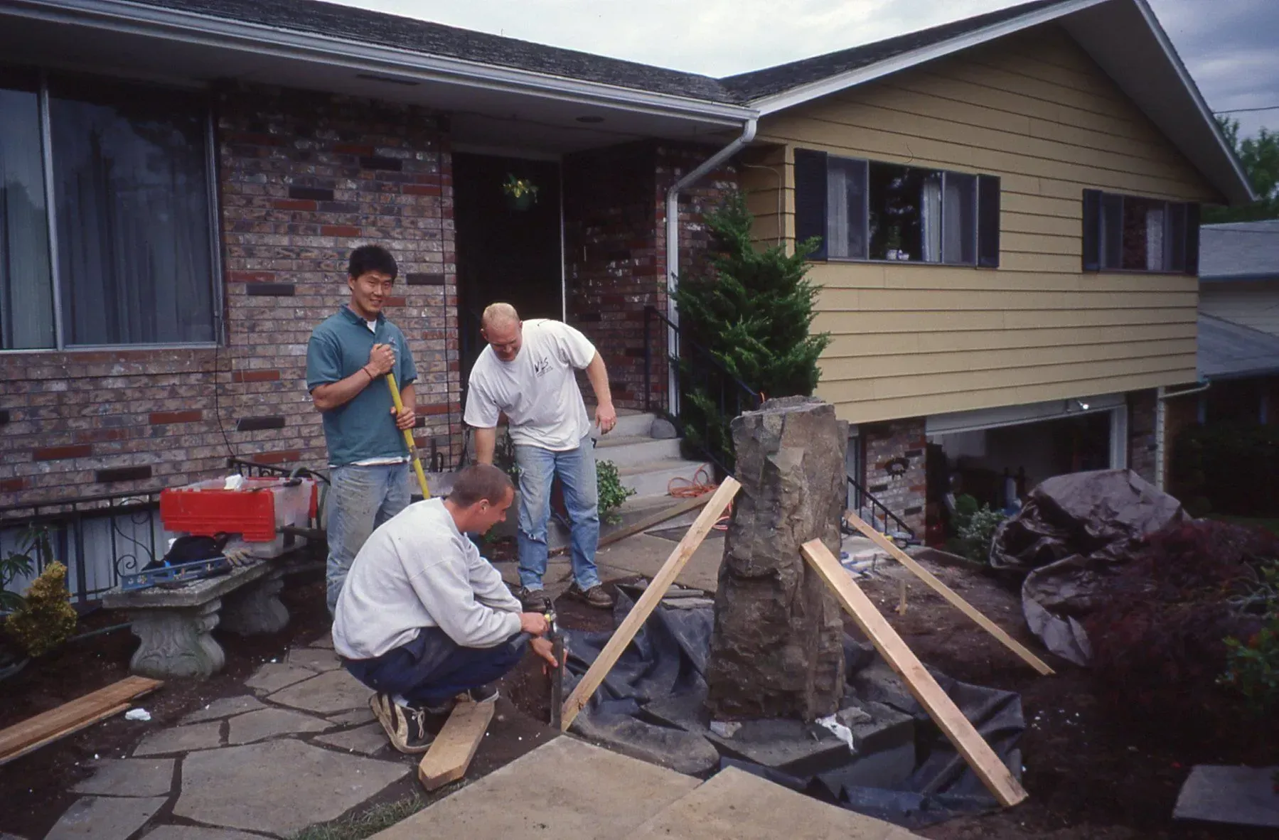 Three men building a fountain in front of a house. One man is squatting, another is shoveling, and the third is looking on.