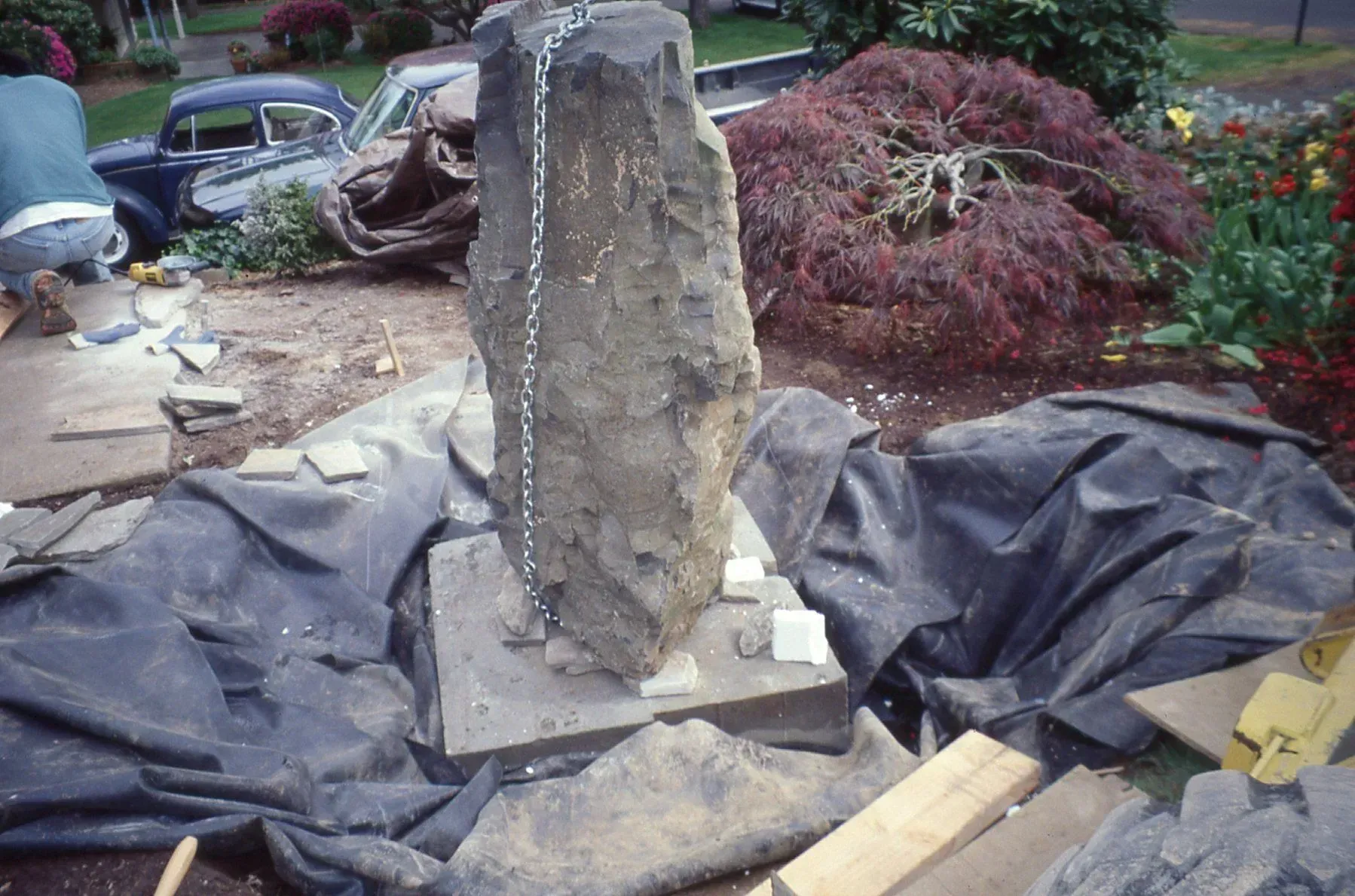 A tall, rough stone being installed in a garden bed, secured with a chain. A car and foliage are in the background.