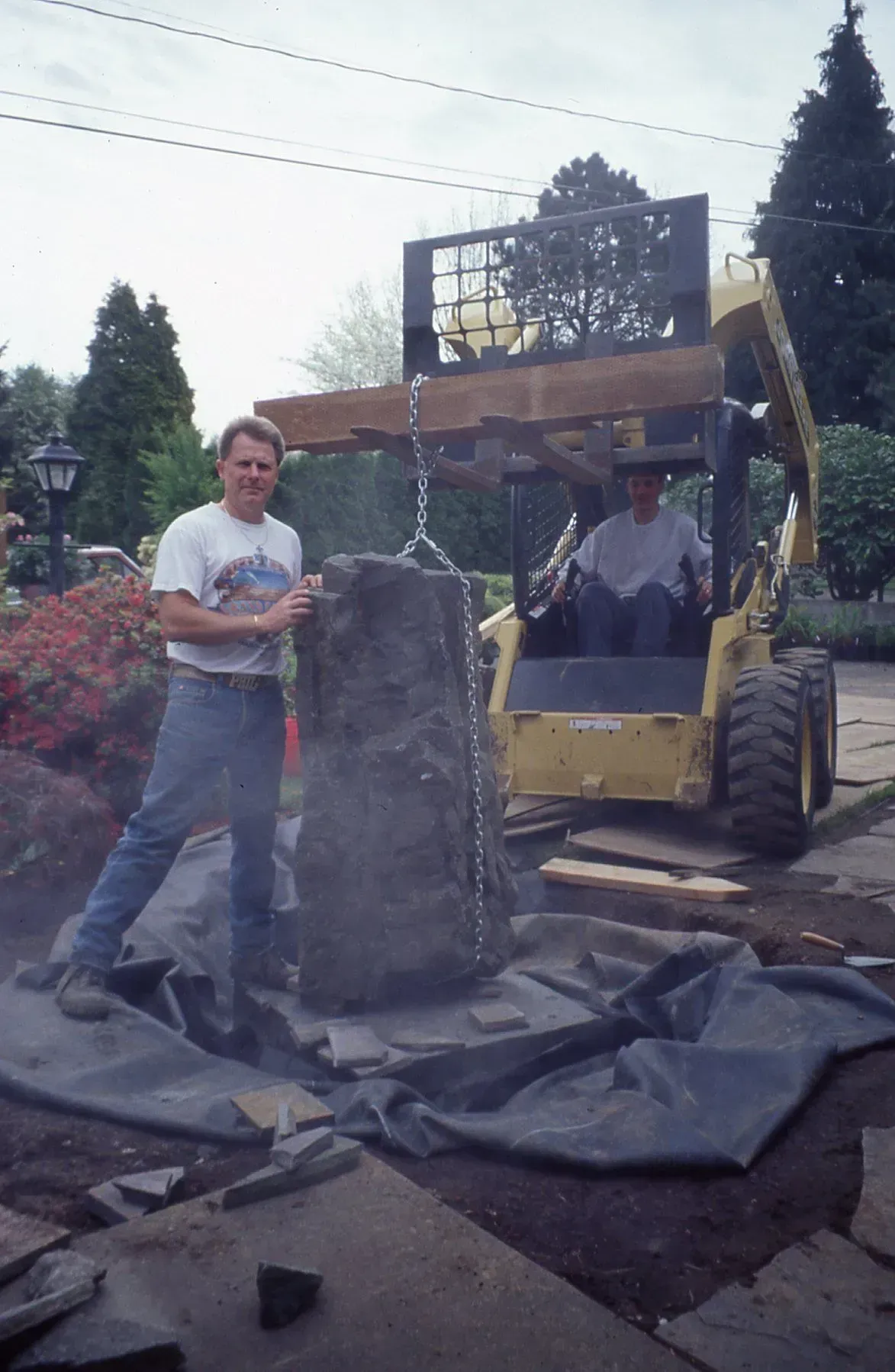Man and forklift lift large rock for landscaping. Another man is in the forklift. They are on a construction site.