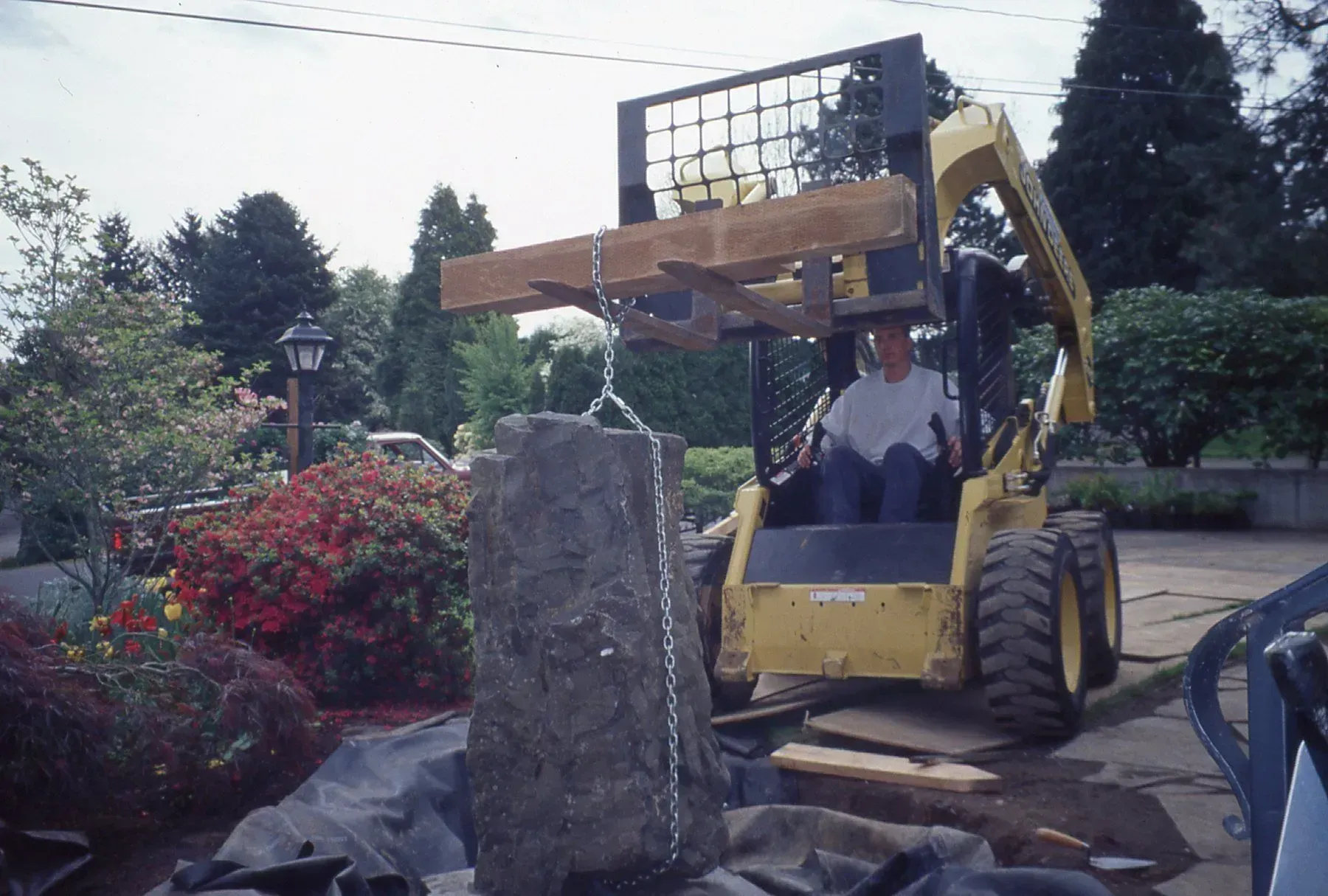 A yellow skid steer lifts a large stone, with a person inside, in front of a garden area.