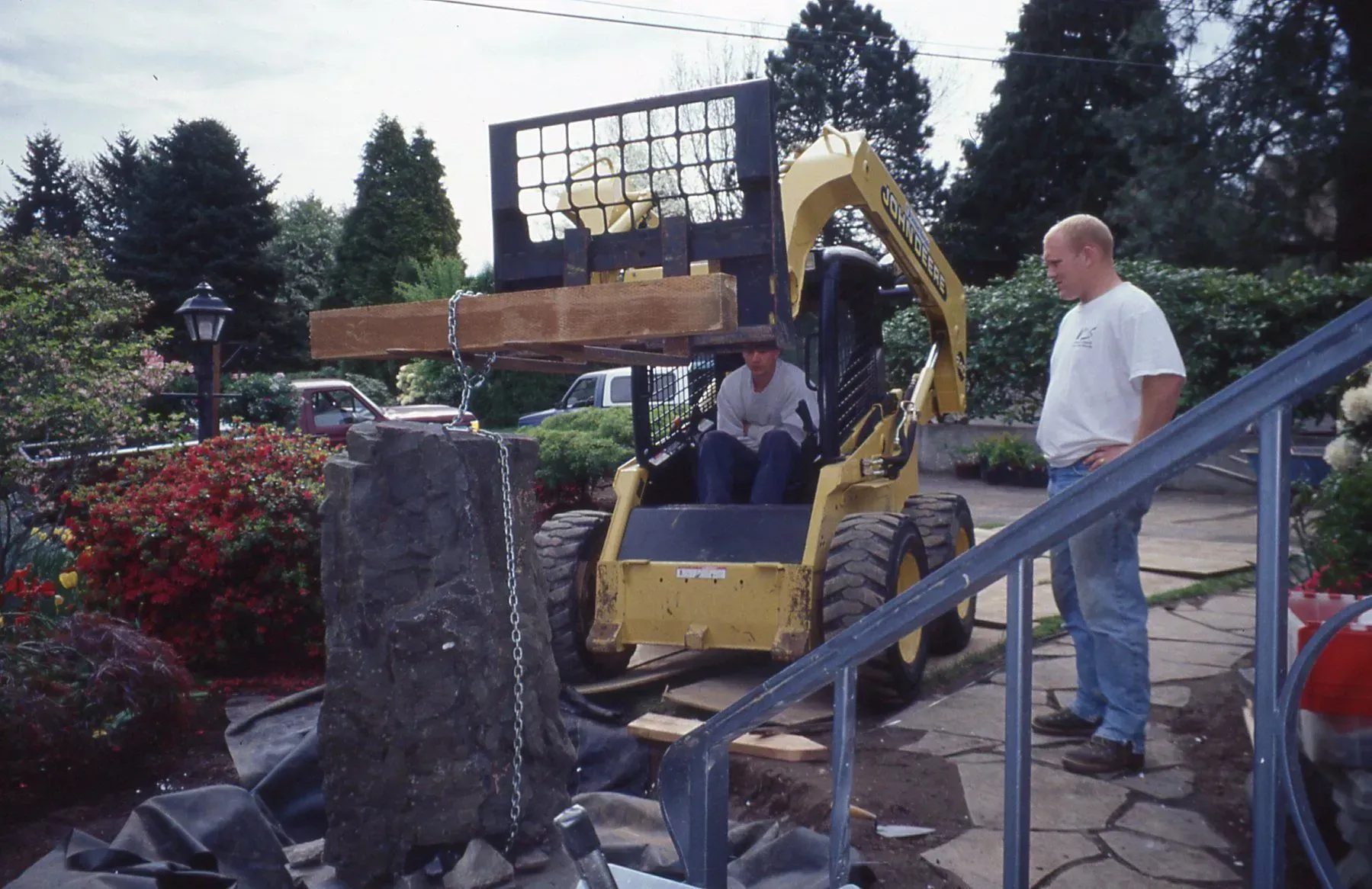 A skid steer loader lifts a large stone, two men watch.  The scene is outside, a house and greenery in the background.