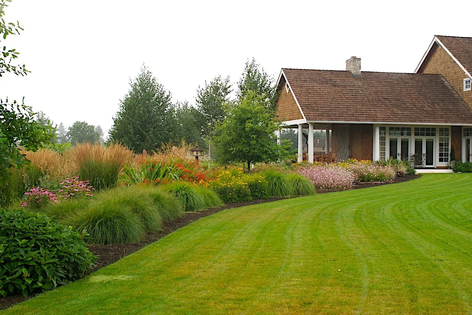 A well-manicured lawn curves past a vibrant flower bed bordering a house with a brown roof and white trim.