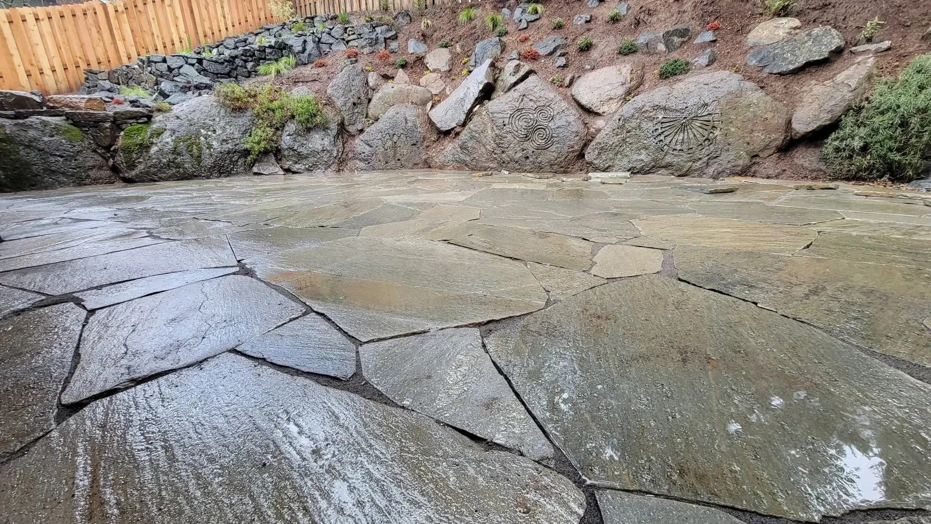Wet stone patio with large irregular flagstones, backed by a retaining wall of large rocks and a wooden fence.