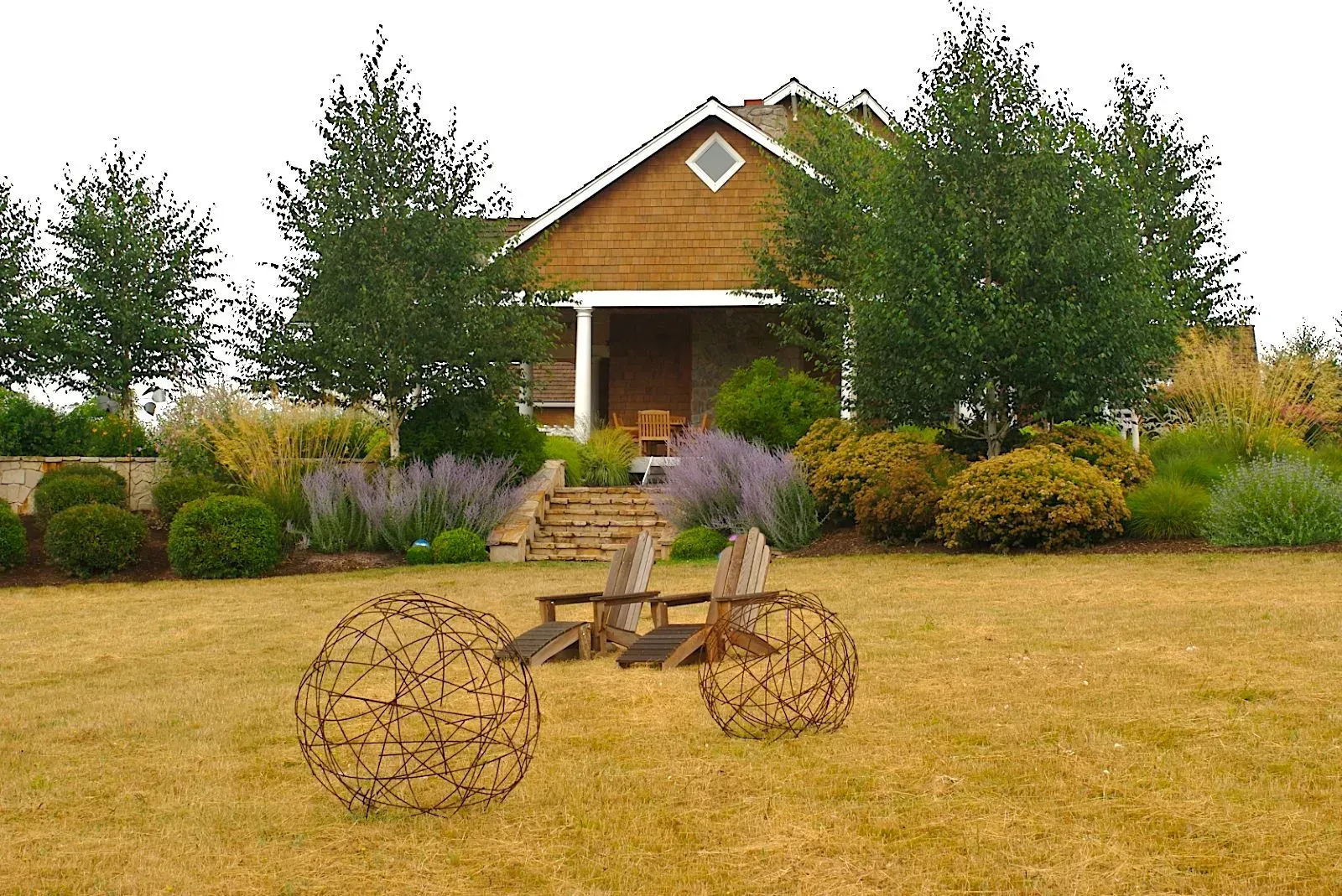 A house with a covered porch is set in a well-manicured garden. Two wire spheres sit on the lawn in front of wooden chairs.