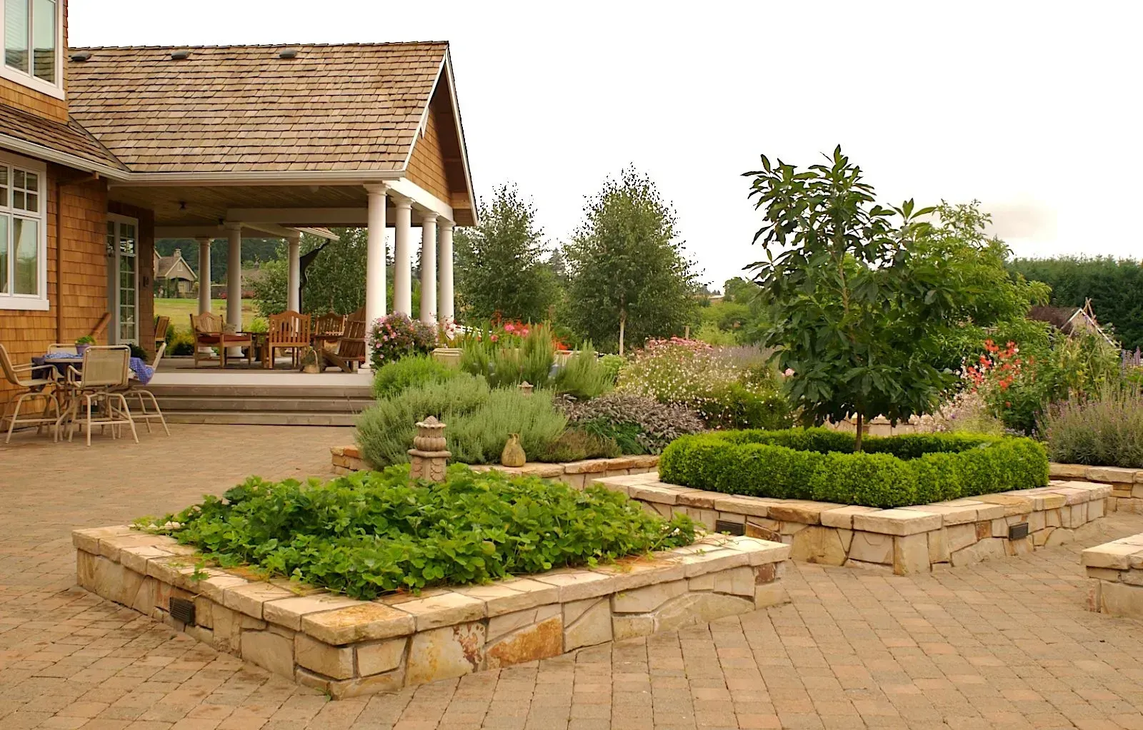 Stone-lined garden beds filled with greenery and a small tree in front of a house with a veranda and a cloudy sky.