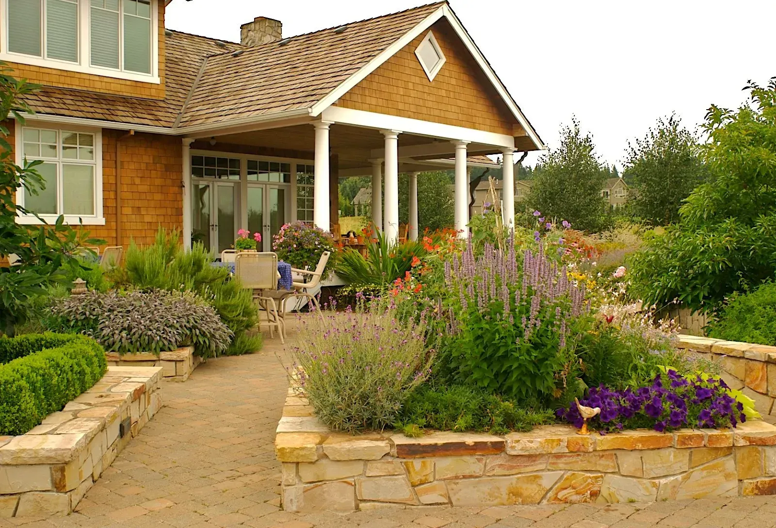 A house with a covered porch, surrounded by stone-lined garden beds filled with colorful flowers and greenery.