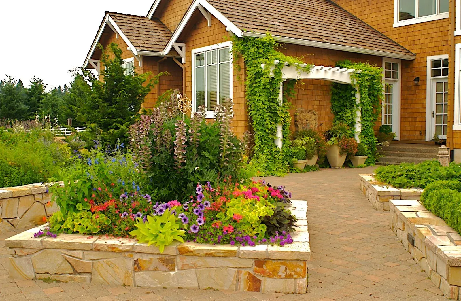 Stone-walled garden bed overflowing with colorful flowers in front of a brown house with ivy-covered pergola.