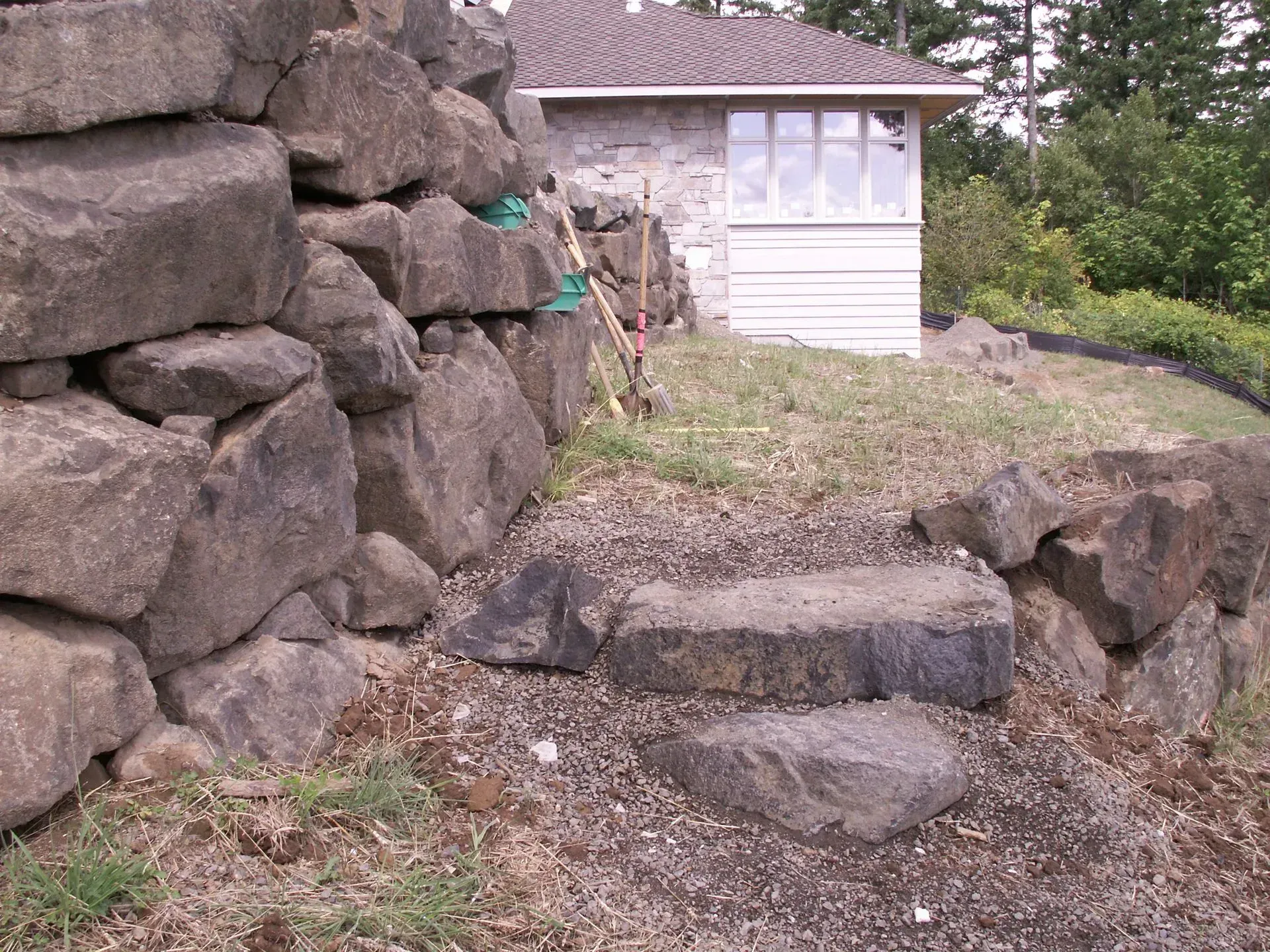 Stone steps lead up a rock wall towards a white building with a brown roof, set in a grassy outdoor area.