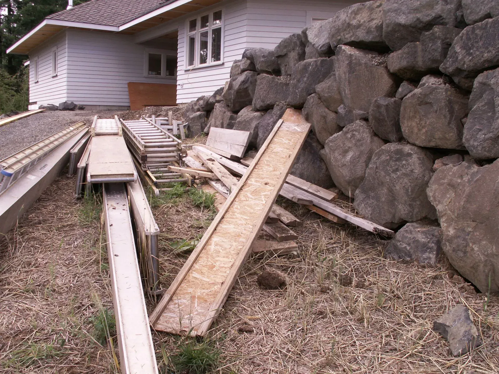 Wooden ramp and construction debris beside a large stone wall, next to a light-colored house.