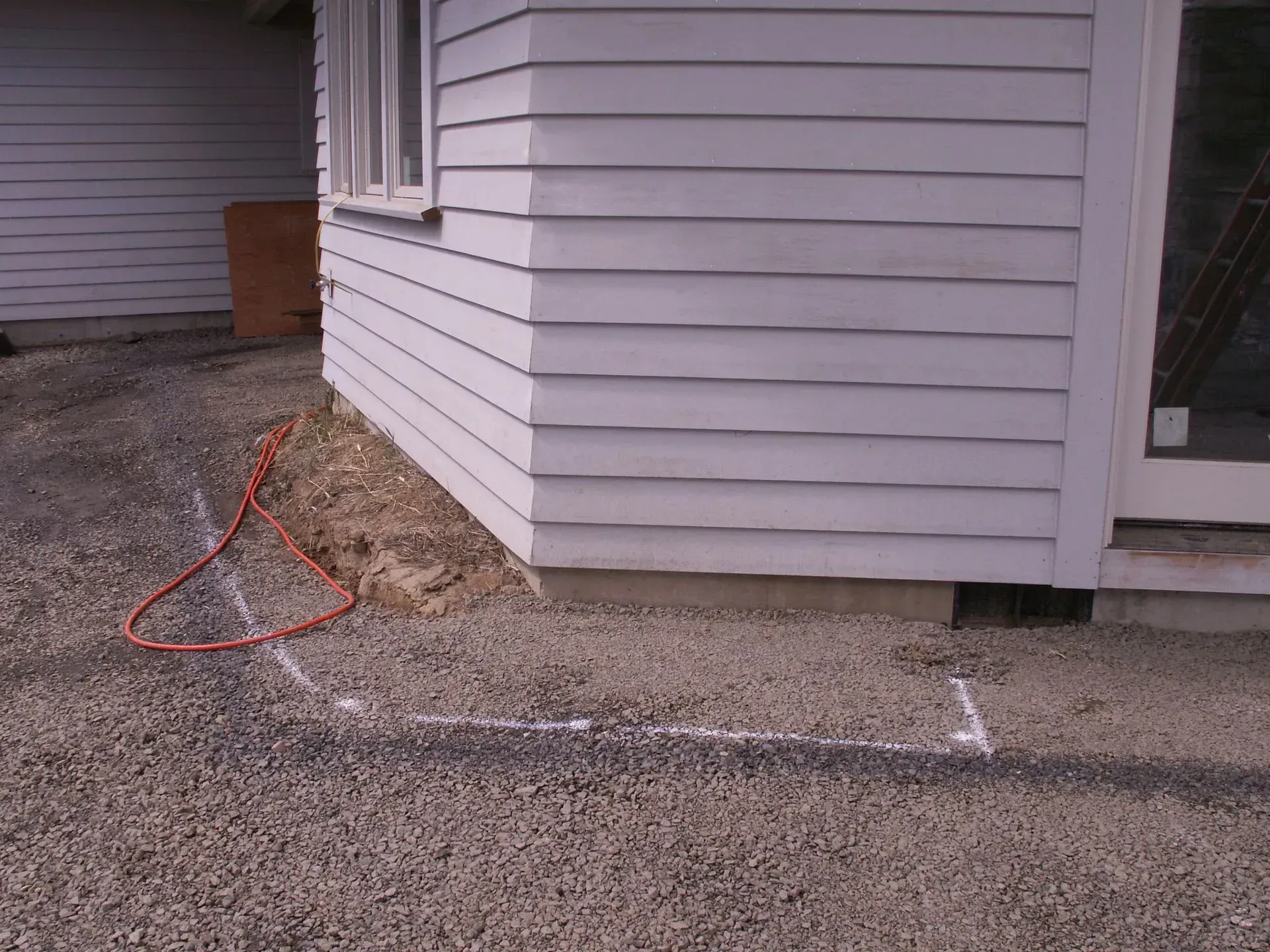 Corner of a white house with a gravel ground. A white chalk outline marks a rectangle in the foreground.