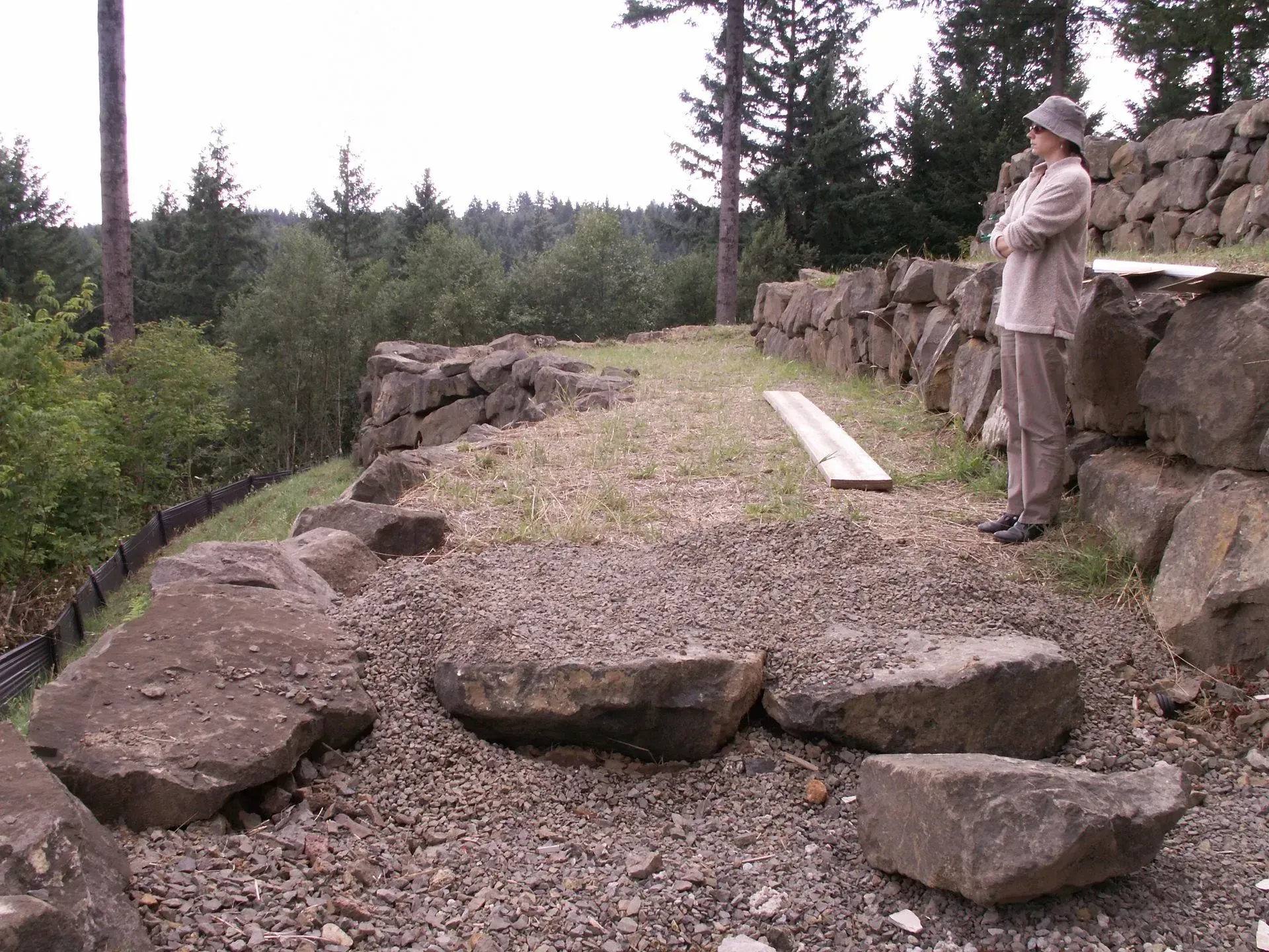 Person observing a construction site with stone retaining walls and a gravel path next to forest.