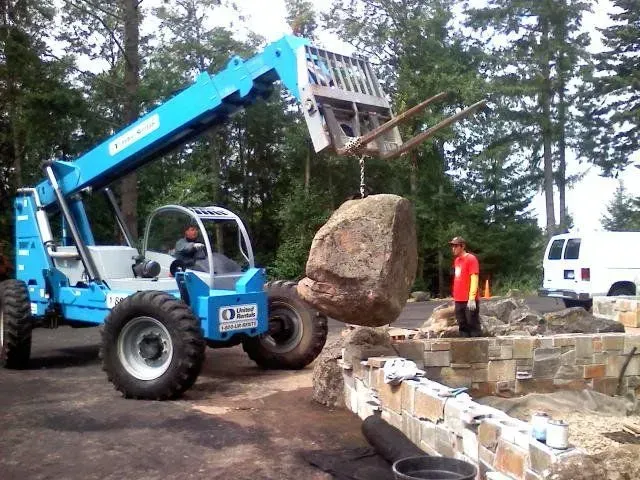 A blue telehandler lifts a large rock at a construction site. A worker in a red shirt watches.