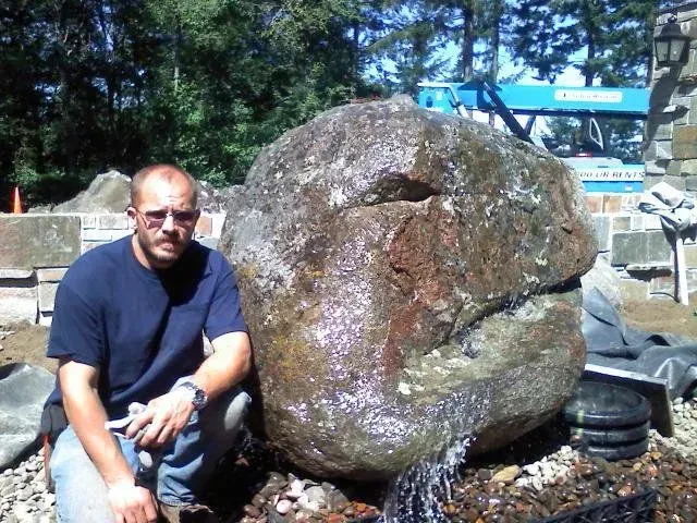 Man kneels next to a large, wet, rock fountain. He wears a blue shirt and jeans. Outdoors.