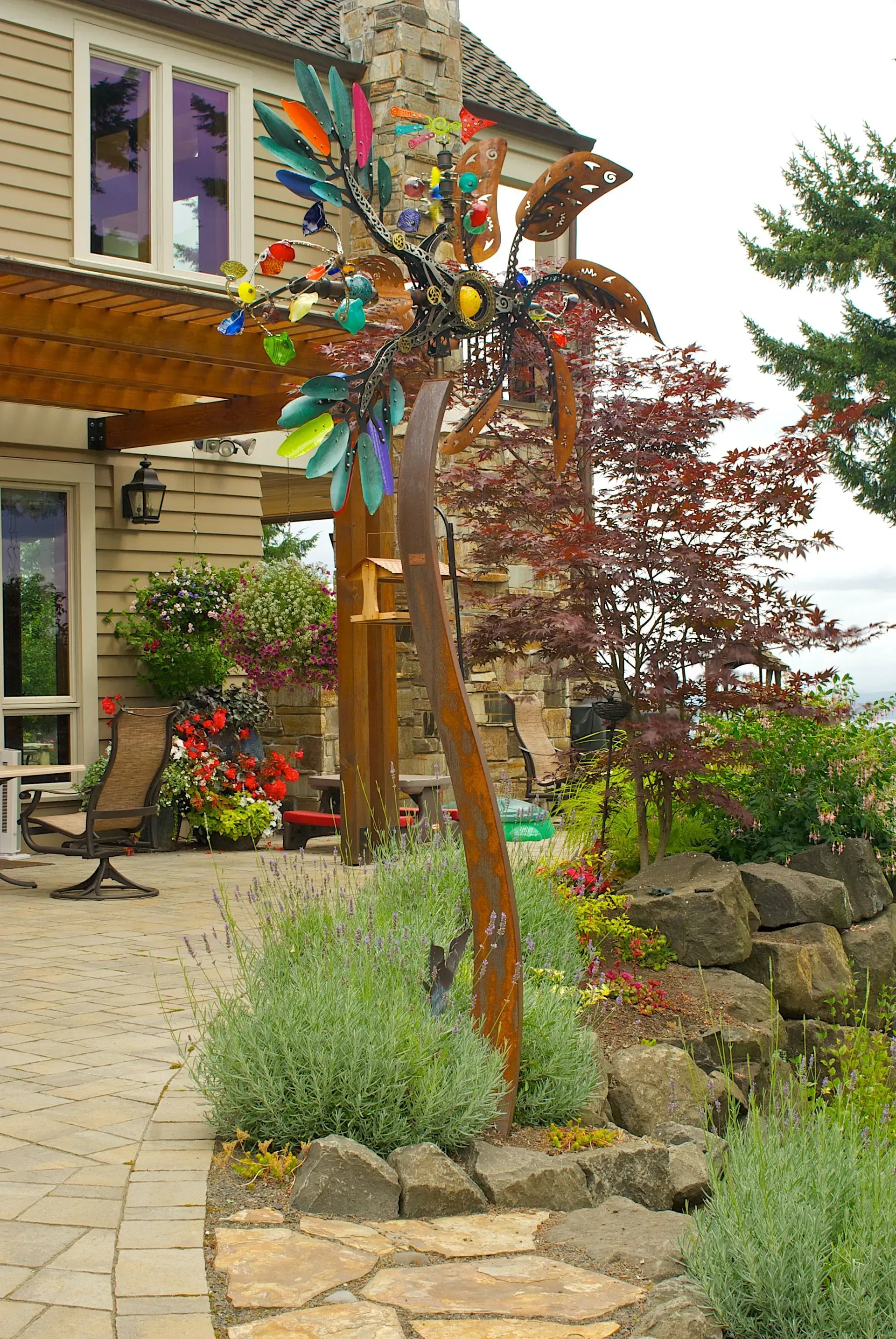 A colorful metal art tree sculpture in a garden setting near a patio.