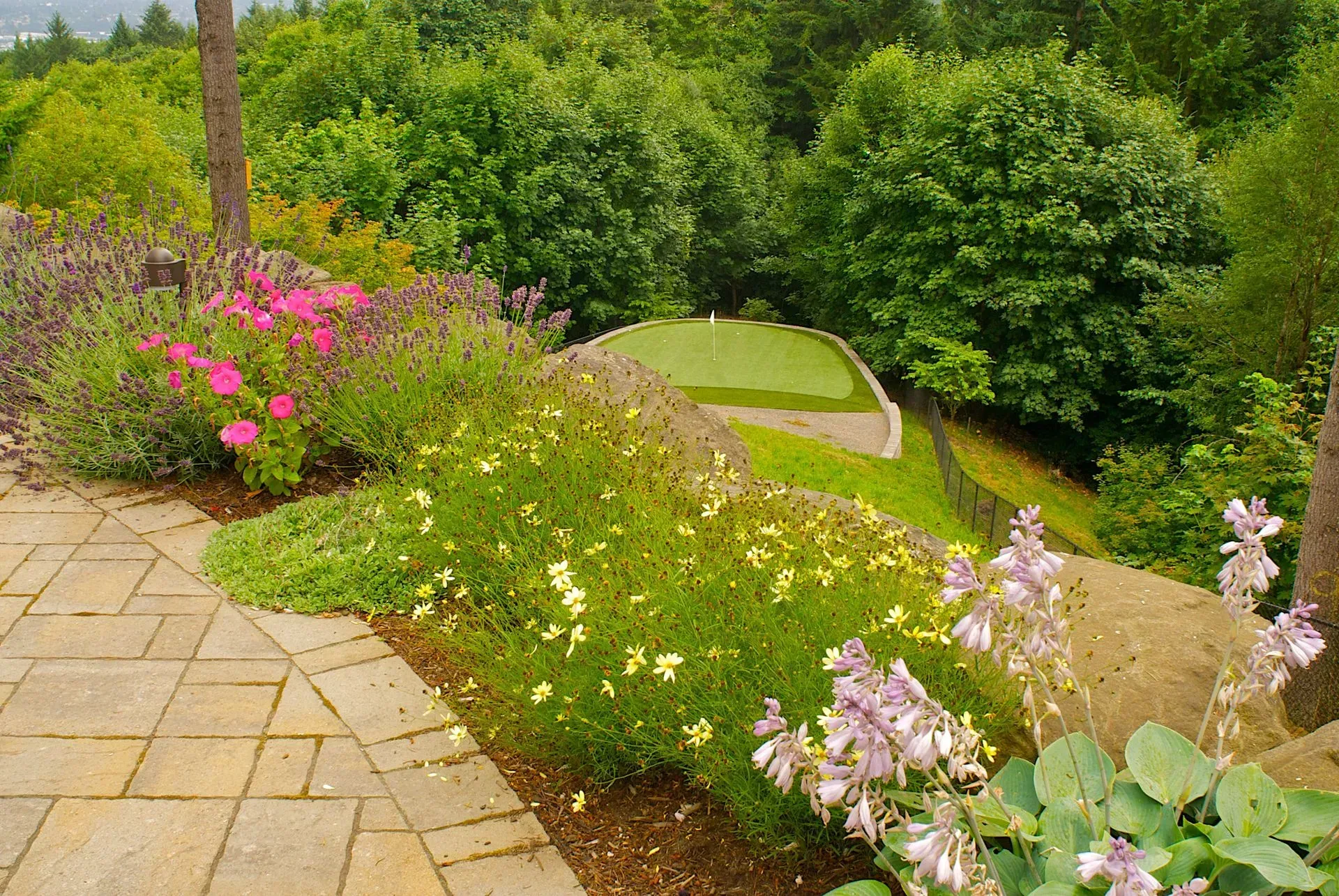 Patio overlooking a lush garden with a putting green. Flowers in pink, purple, and yellow bloom around the rocks and foliage.
